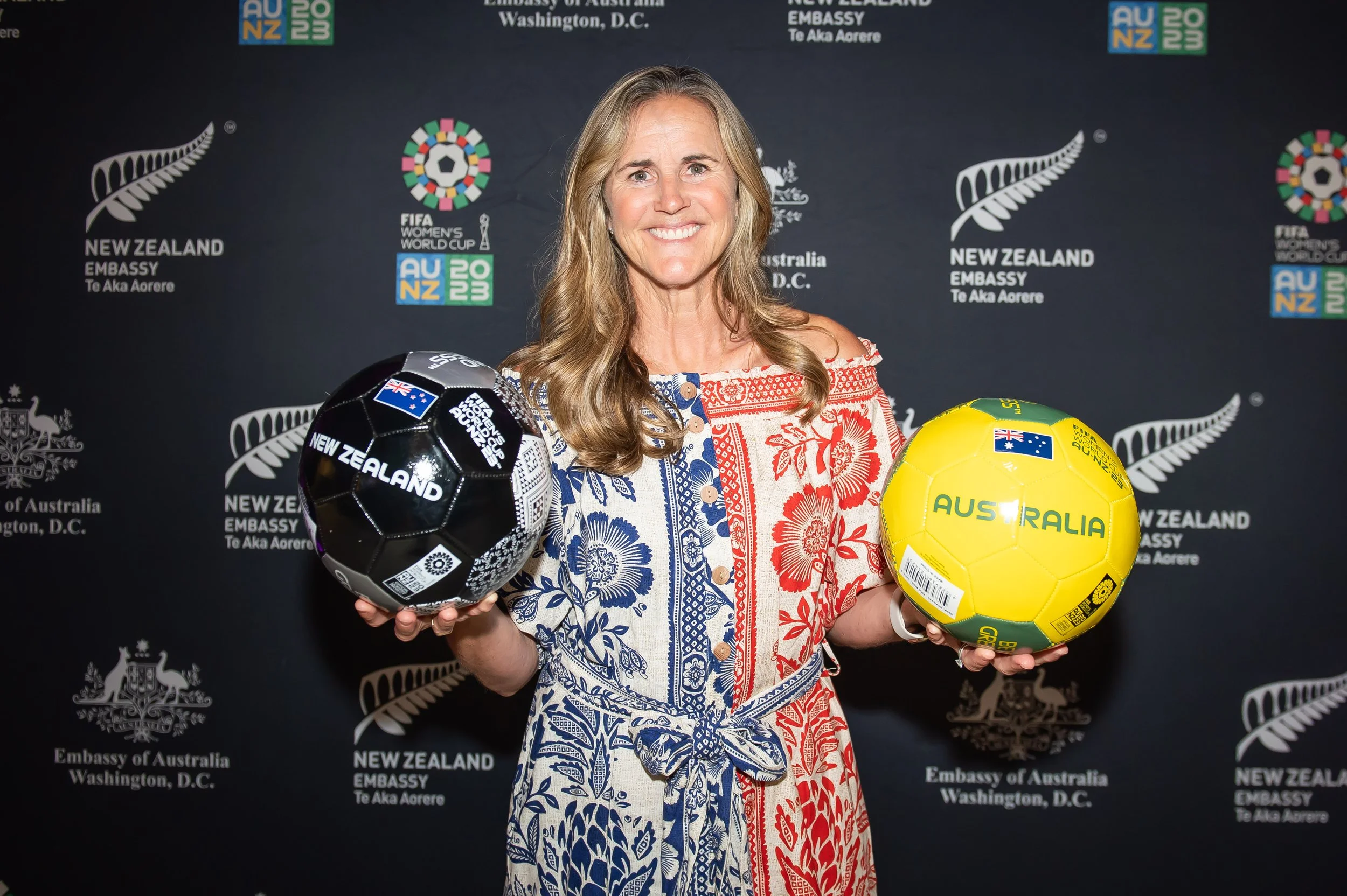 A woman with long wavy blonde hair smiling while holding two Australian national soccer balls, one black and white and the other yellow. She is standing in front of a dark backdrop with logos of New Zealand Embassy and FIFA Women's World Cup.