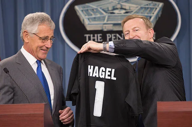 Two men, one in a suit and one in a dark jacket, holding a black sports jersey with the name 'HAGEL' and the number 1, in front of a sign that reads 'THE PENTAGON'.