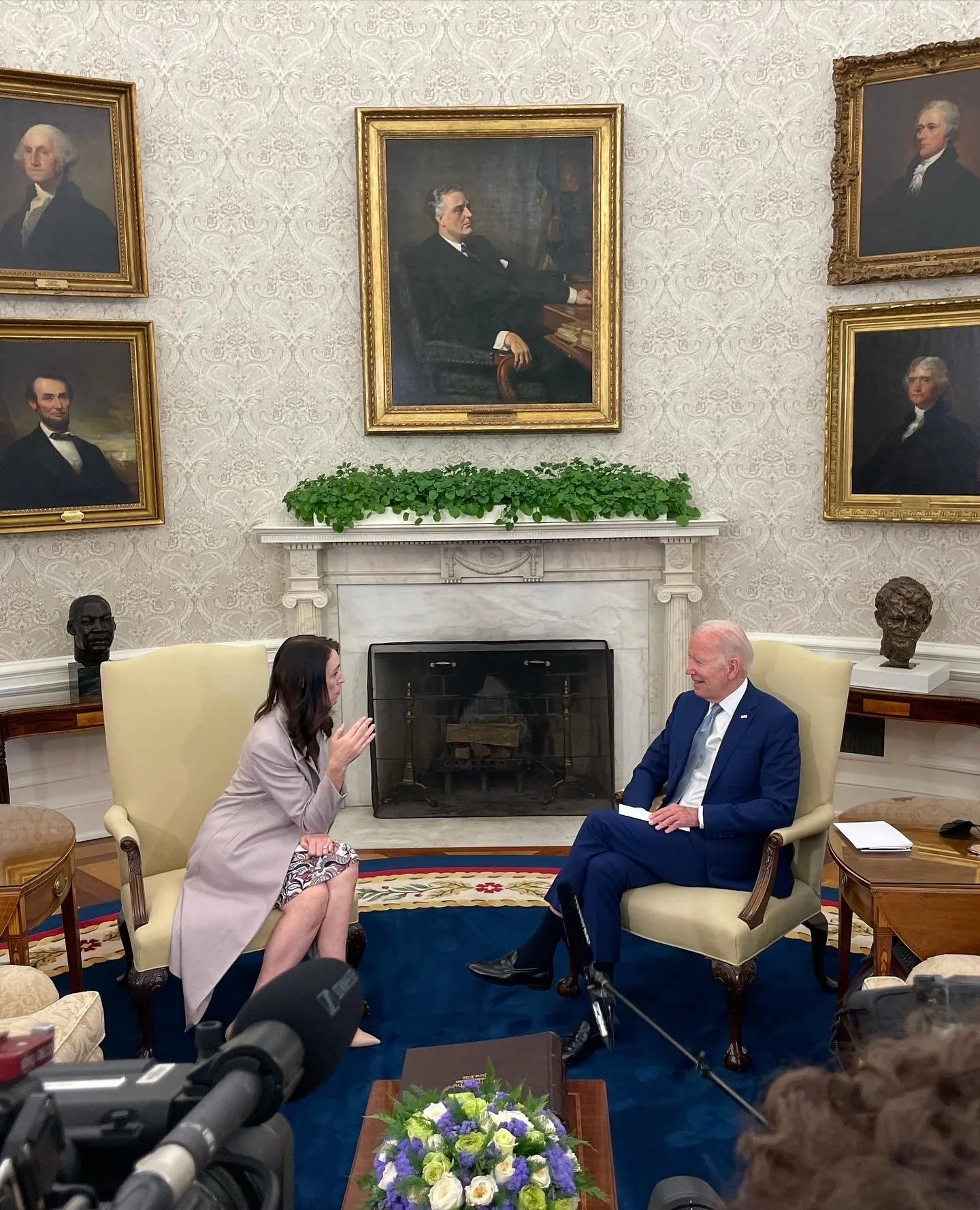 President Joe Biden and a woman engaged in conversation in a formal room with portraits and busts on the wall, and a fireplace at the center.