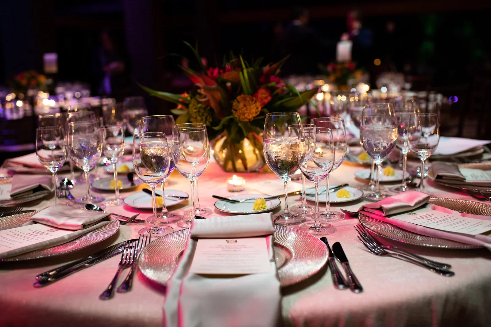 Elegant banquet table setting with multiple wine glasses, a floral centerpiece, and place settings with menus, silverware, and napkins, illuminated with pink and purple lighting.