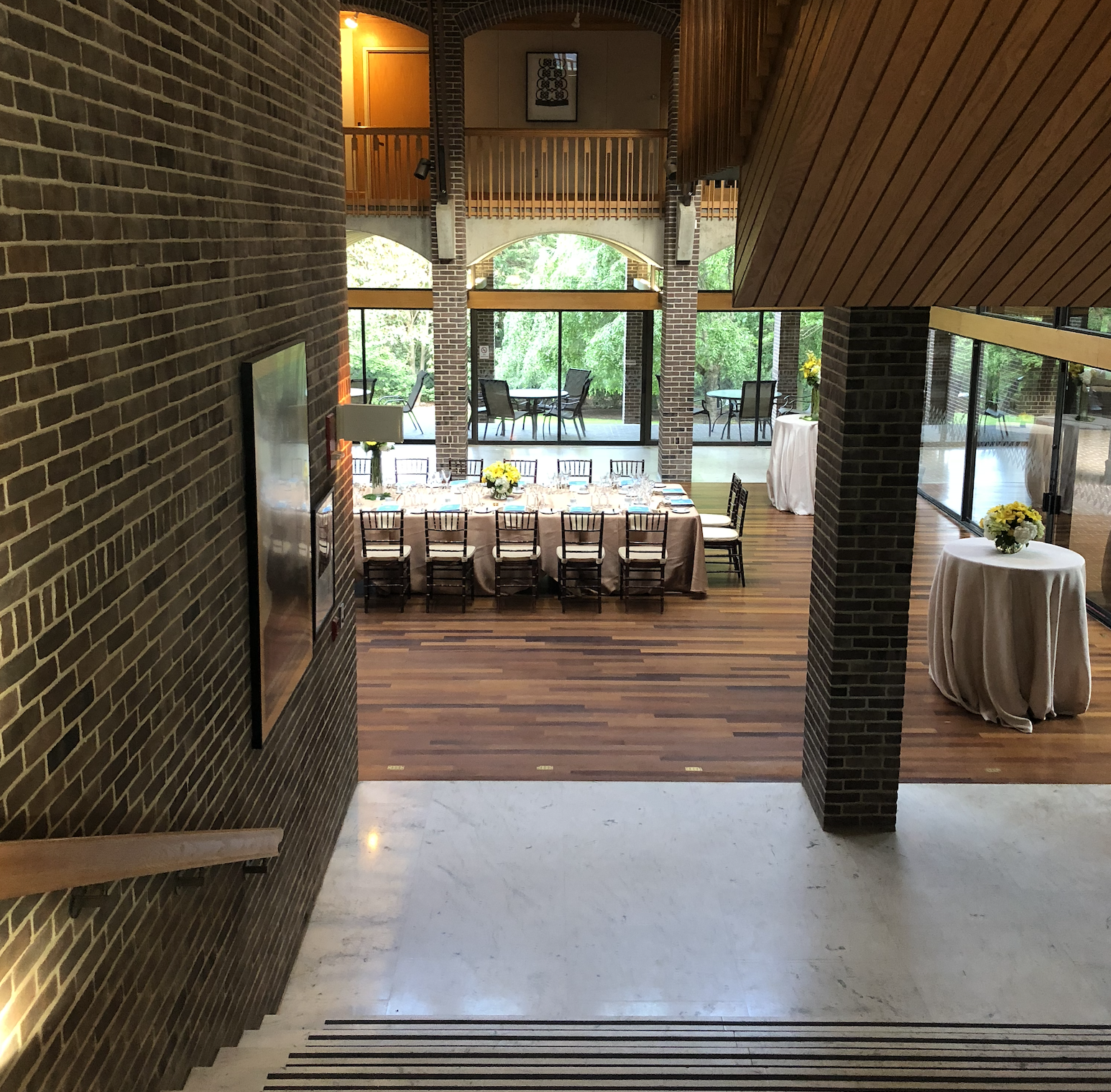 Interior view of a dining room with a long table set for a formal meal, decorated with yellow and white flower arrangements. The room features large windows with outdoor views, wooden flooring, brick and wood-paneled walls, and additional seating on a balcony. Two round tables with flower centerpieces are positioned to the right.