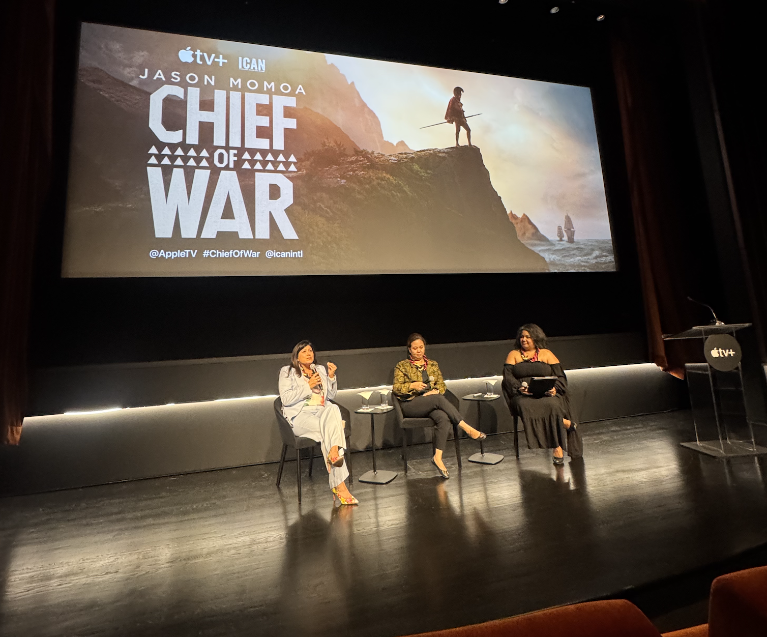 Three women sit on stage in front of a large screen displaying promotional poster for 'Chief of War,' including the Apple TV+ logo and an image of a person standing on a cliff with two ships in the distance. The women are engaged in discussion, with one speaking into a microphone.