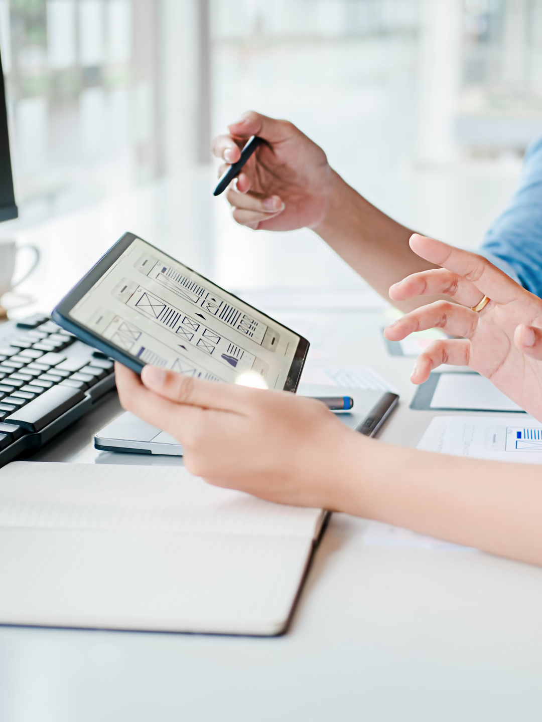 Two people working at a desk, one holding a tablet with wireframe sketches, the other gesturing with a pen.