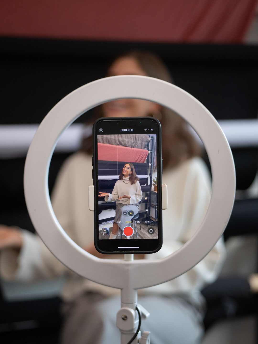 A woman sitting on a stool, speaking and gesturing with her hands, is reflected in a ring light while recording a video with her phone.