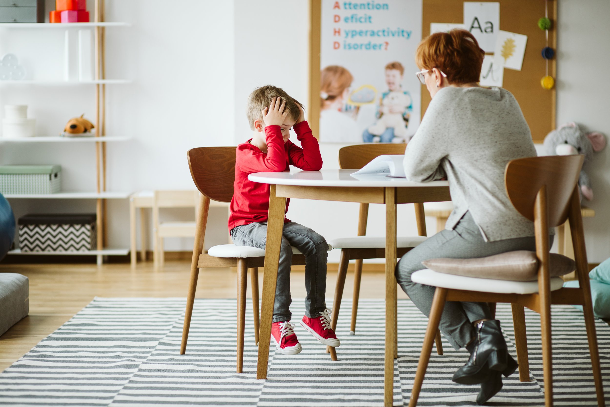 A young boy with tousled hair, wearing a red long-sleeve shirt, gray jeans, and red sneakers, sits at a white table with his hands on his head, looking distressed. An adult woman, possibly a therapist or teacher, with short reddish hair, glasses, a light gray sweater, and dark pants, sits across the table facing him. The room has a striped rug, shelves with toys and books, and a bulletin board with educational posters and decorations.
