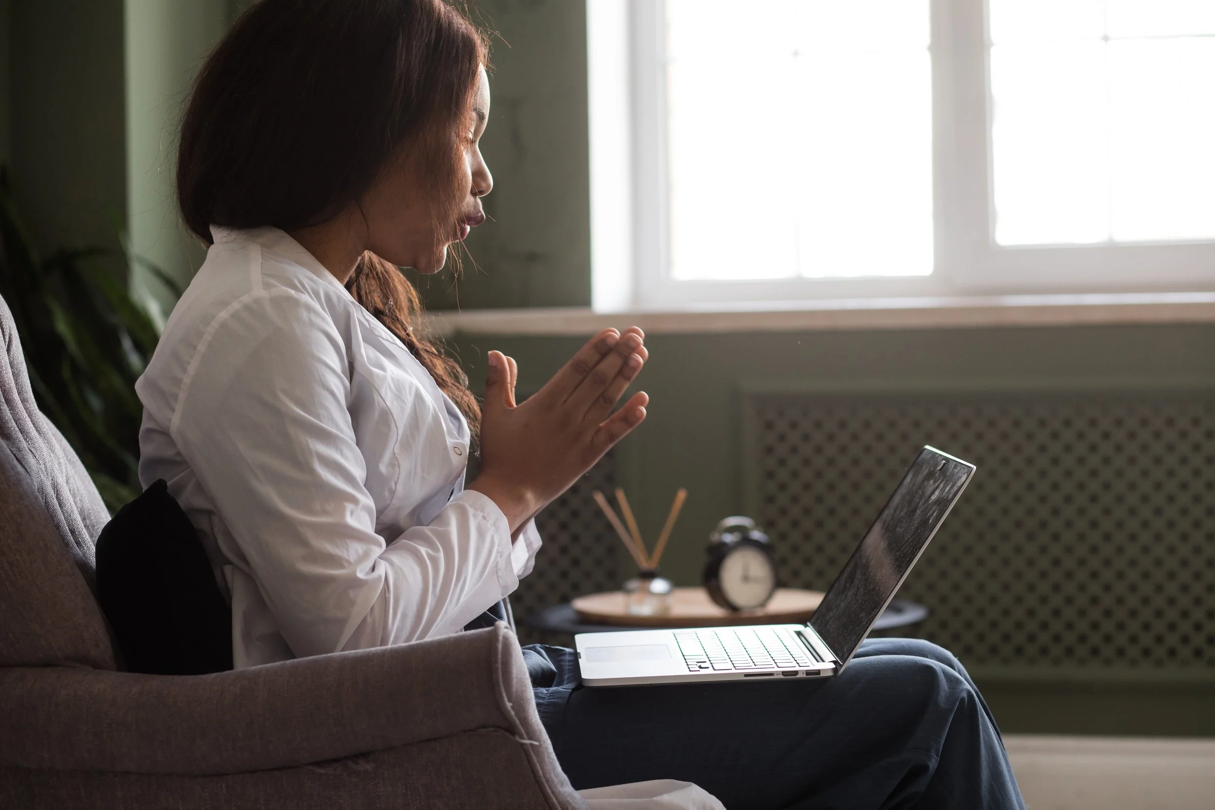 A woman sitting on a gray sofa, wearing a white shirt and black pants, with her hands raised in a prayer position. She is using a laptop on her lap, and there is a clock and some small decorative items on the table behind her. The room has a large window in the background.