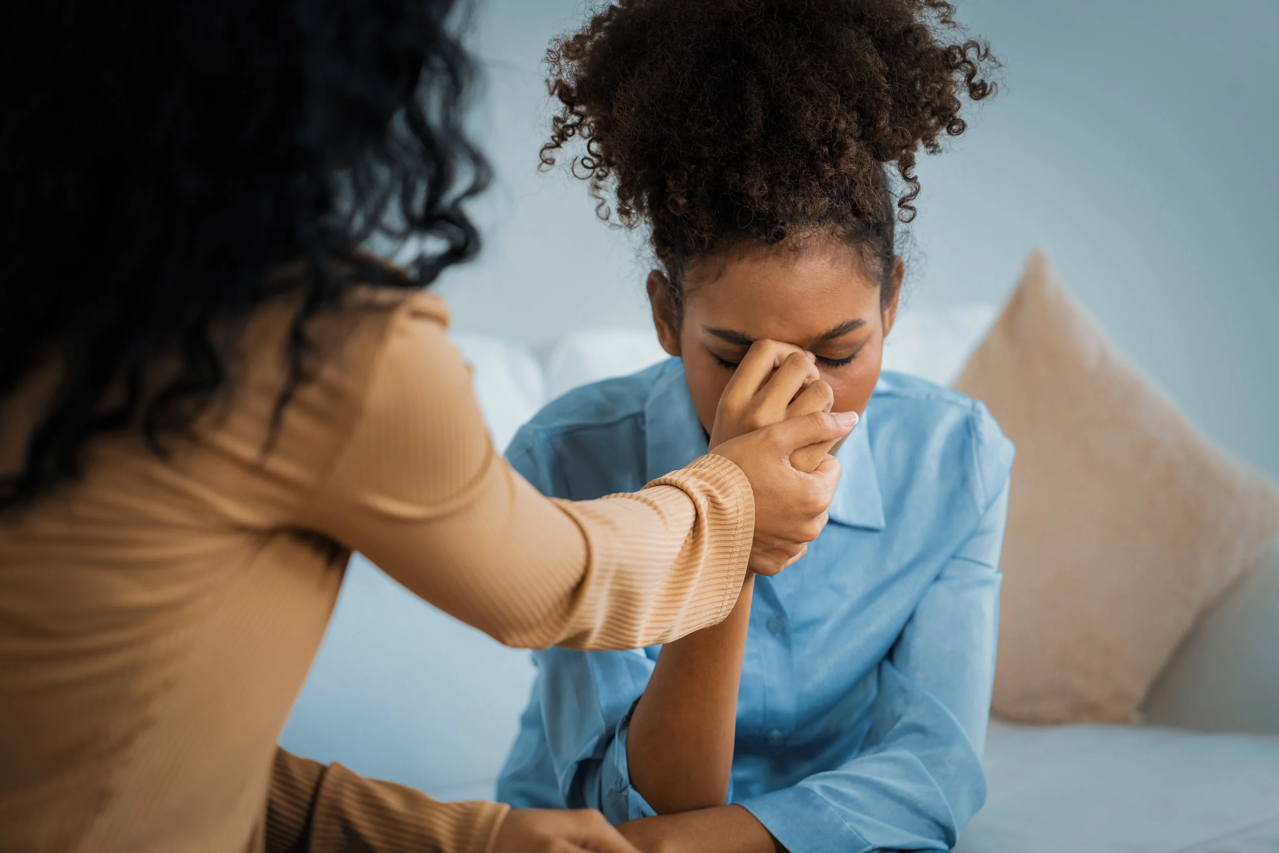 Two women engaged in a comforting moment, one woman is holding the hand of the other woman who appears distressed and is pinching the bridge of her nose while closing her eyes.