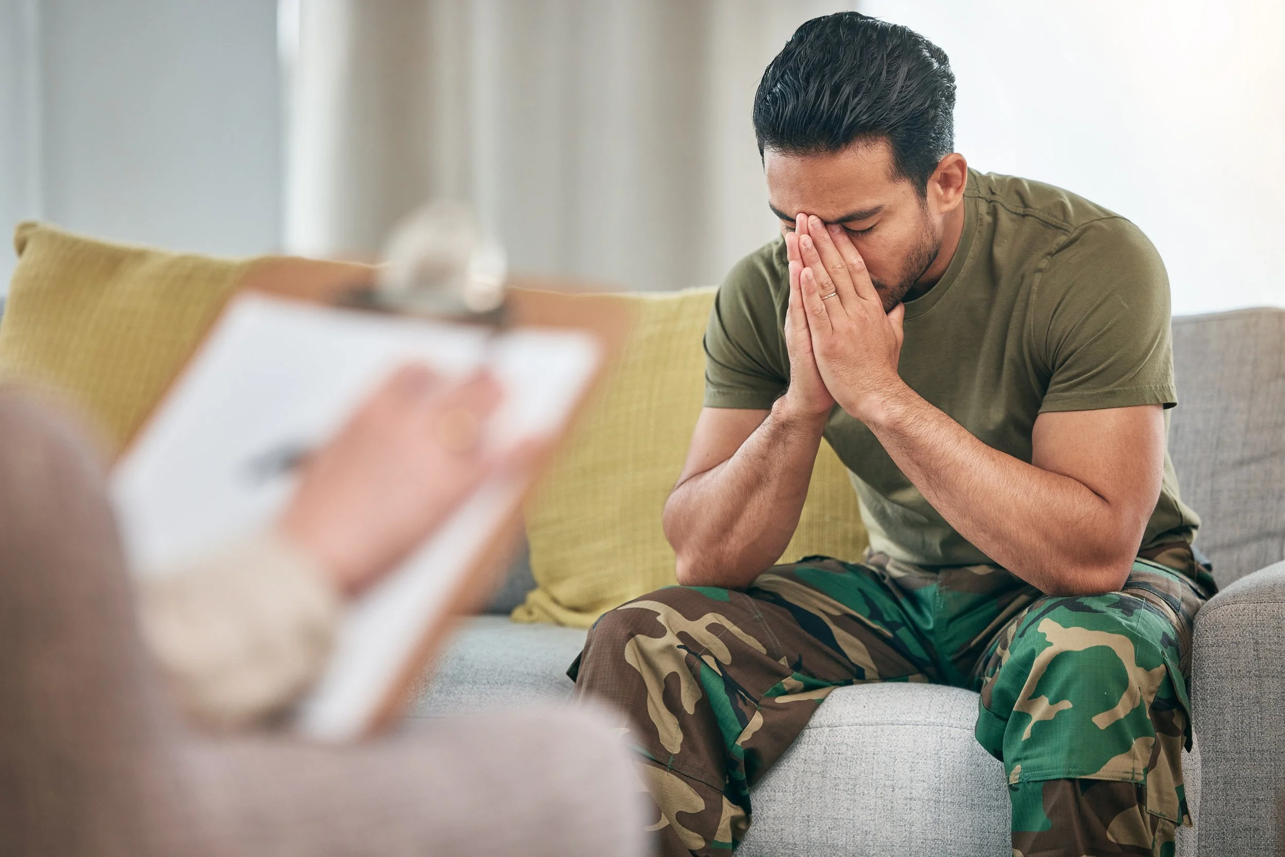 A man in military camouflage attire kneeling on a couch with his hands clasped in front of his face, appearing distressed or upset.