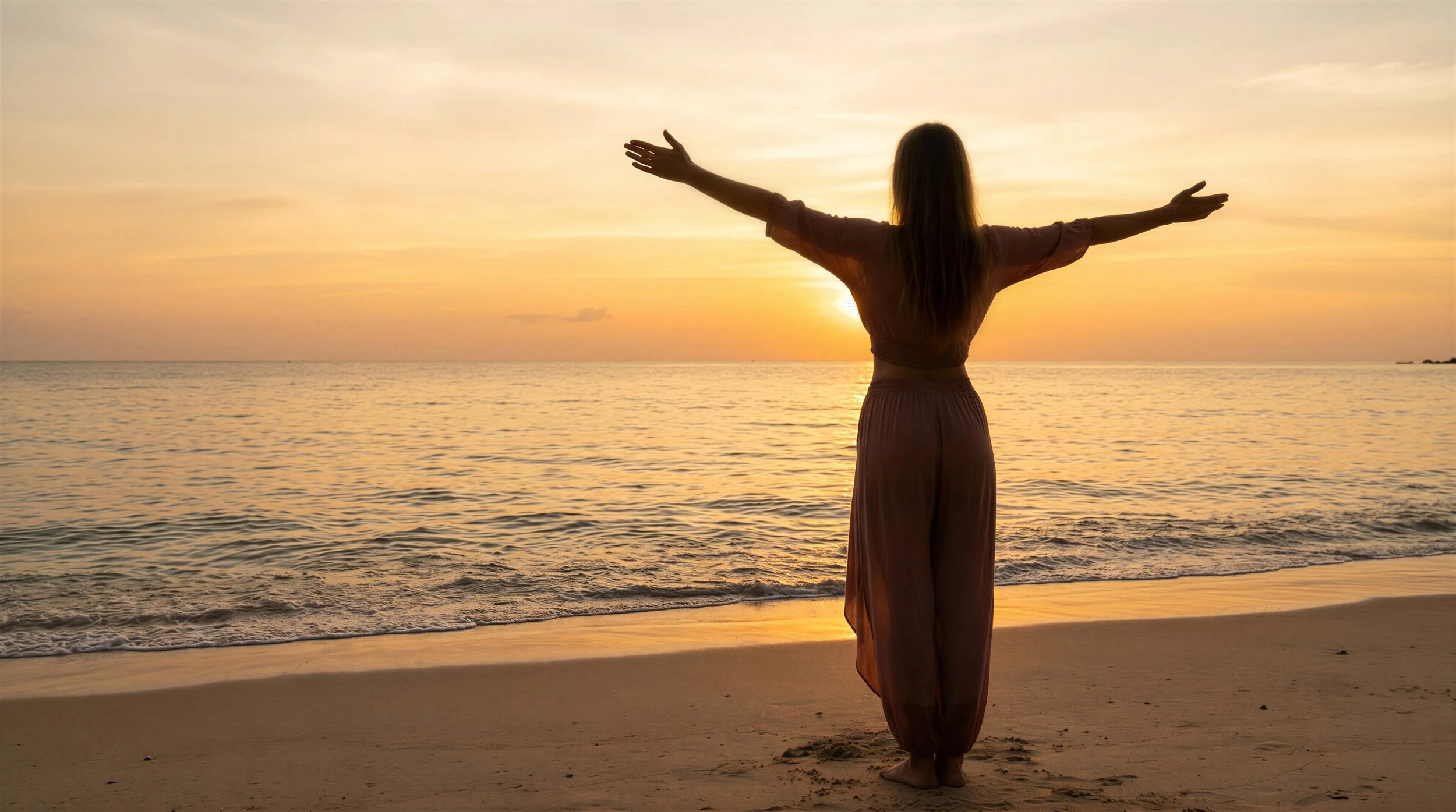 A woman standing barefoot on the beach with arms outstretched, facing the ocean and sunset, wearing a long dress.