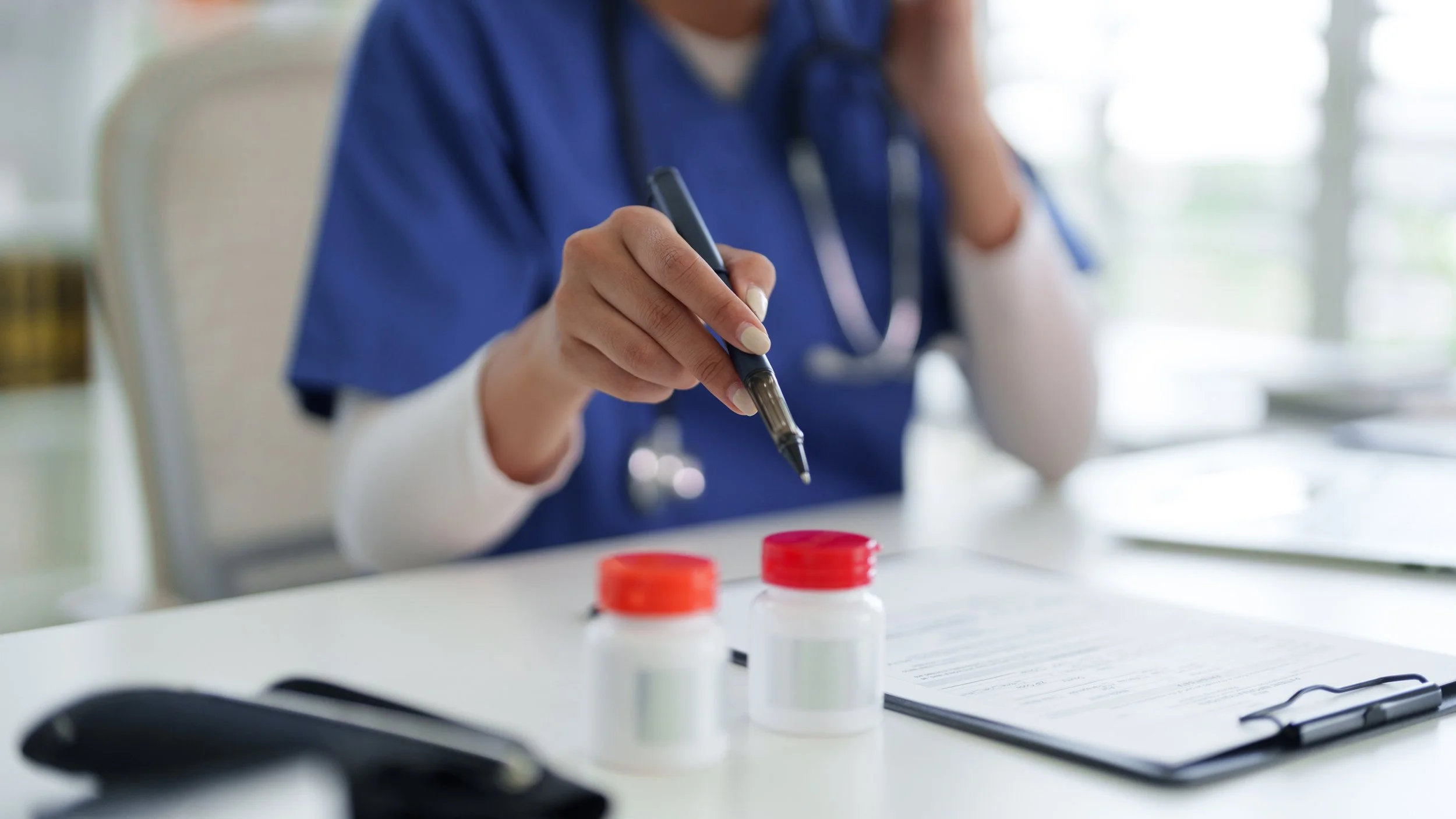 A healthcare worker in blue scrubs is holding a syringe over a table with medicine bottles and a clipboard, in a bright medical setting.