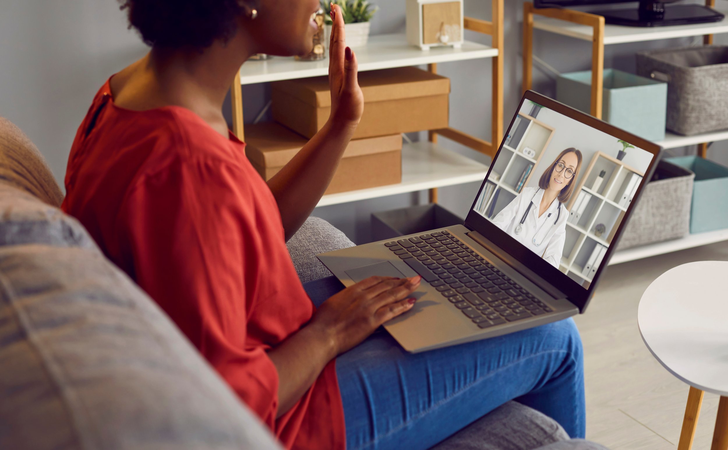 A woman in a red shirt sitting on a gray couch engaging in a video call with a female doctor on her laptop in a room with shelves and storage boxes.