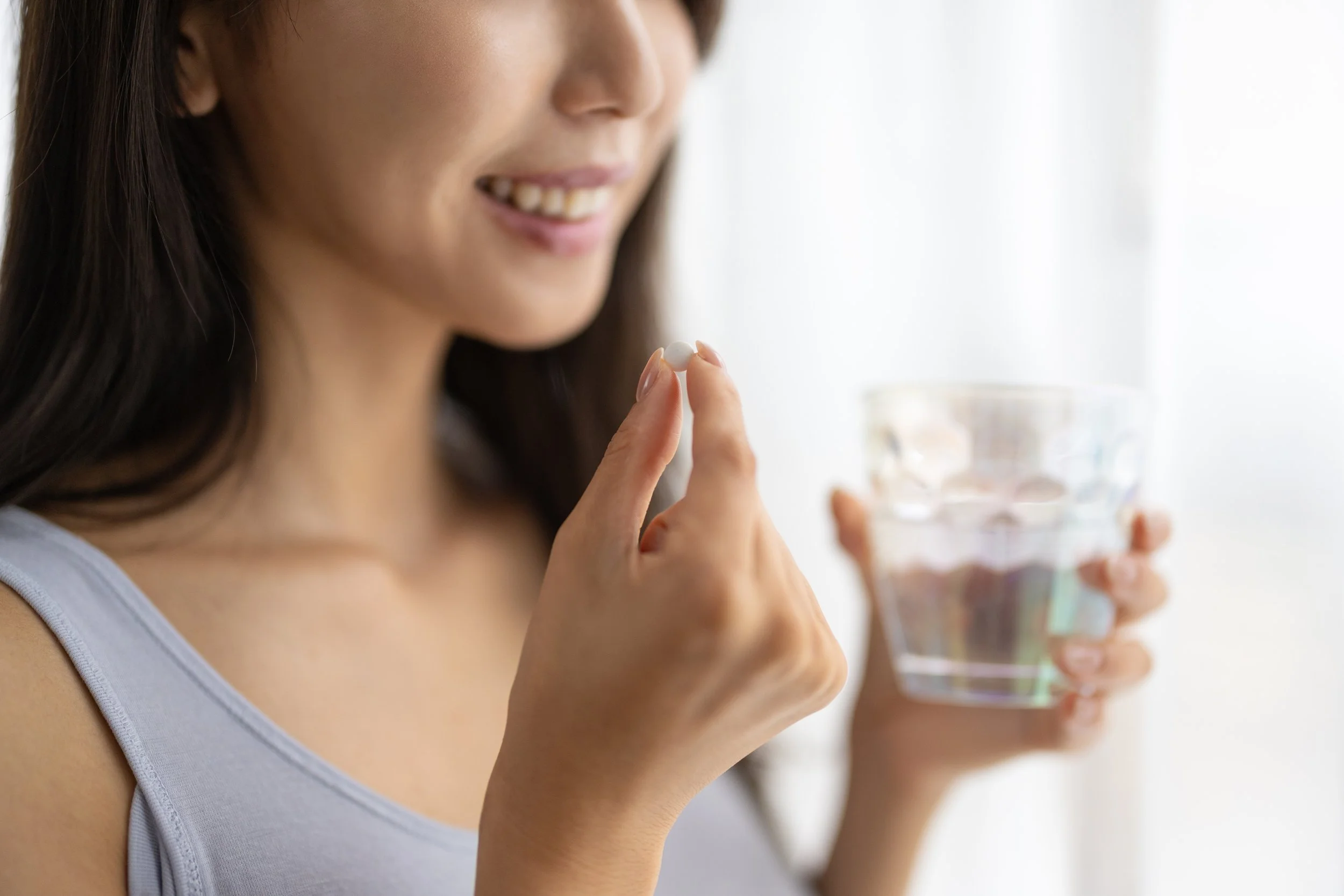 A woman smiling and holding a pill in her finger with a glass of water in the background.