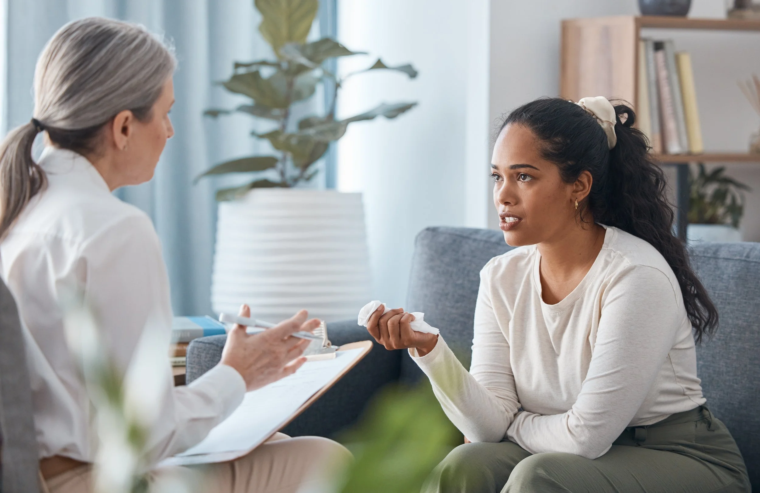 A woman with dark hair tied with a cream-colored scrunchie is sitting on a gray couch and talking to an older woman with gray hair tied in a ponytail. The conversation appears serious. The background shows a bookshelf and a plant, and the setting looks like a living room.