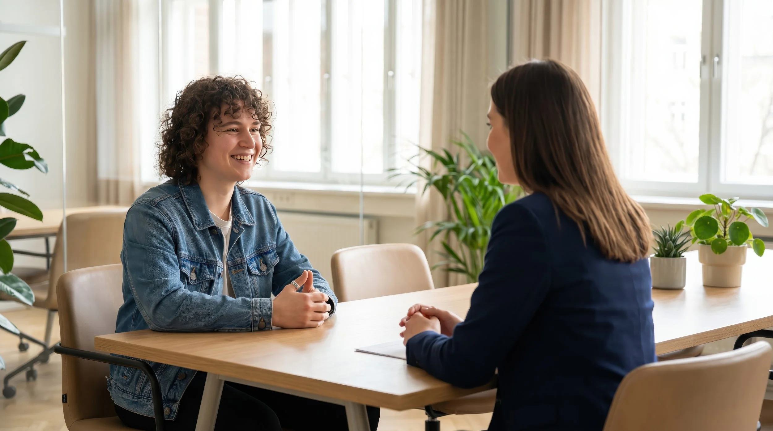 Two women having a conversation at a table in a bright room with large windows and green plants.