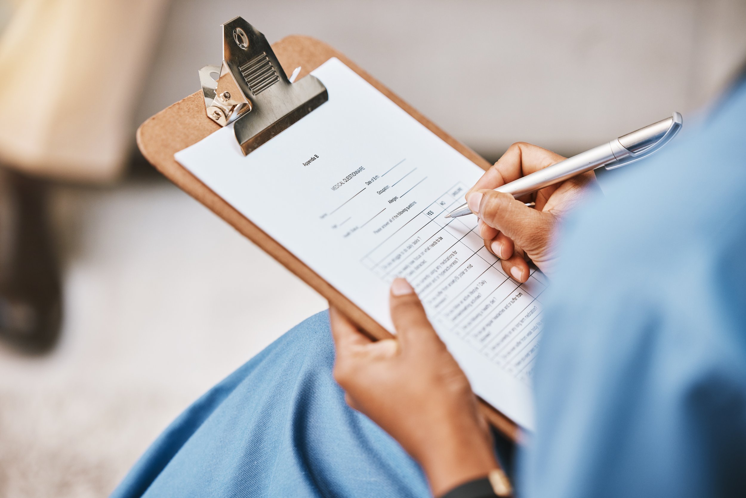 Person writing on a clipboard with a medical questionnaire, with a metal clipboard clip and a silver pen.