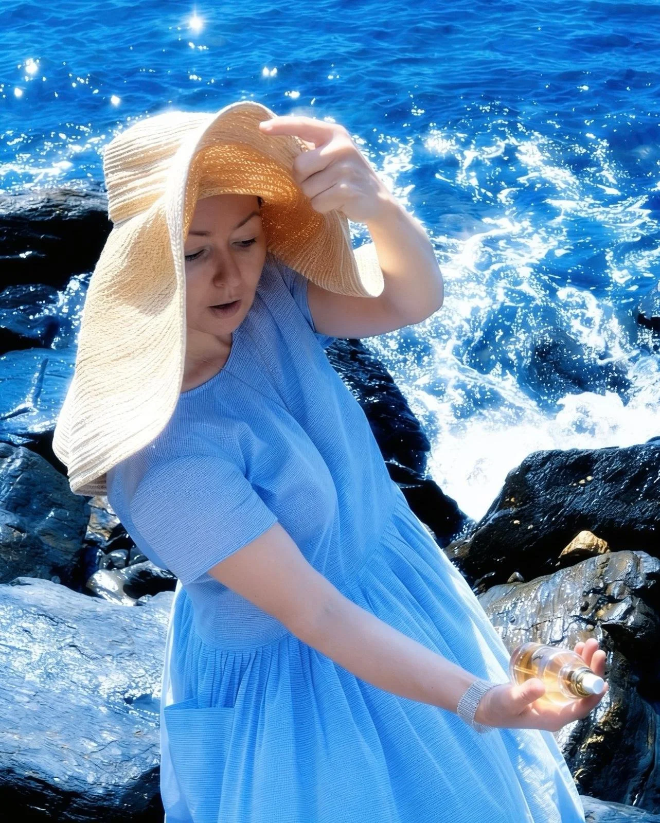 A woman wearing a large straw sunhat and blue dress stands on a rocky shoreline with ocean waves in the background, holding a small bottle of perfume.