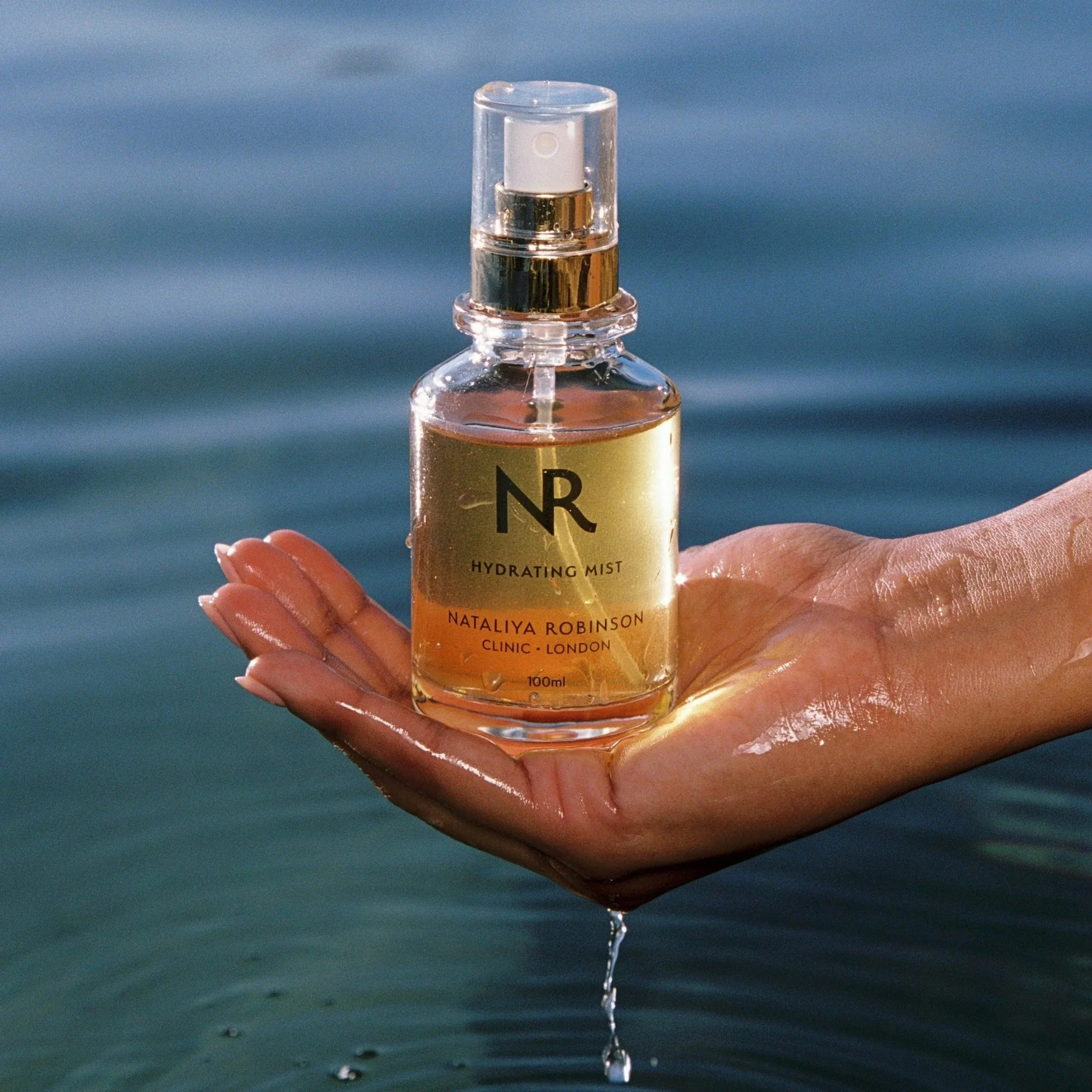A clear glass spray bottle labeled 'Hydrating Mist' by Nataliya Robinson, held by a person with neatly manicured nails, with water droplets dripping from the bottle against a blurred blue-green background, likely near water.