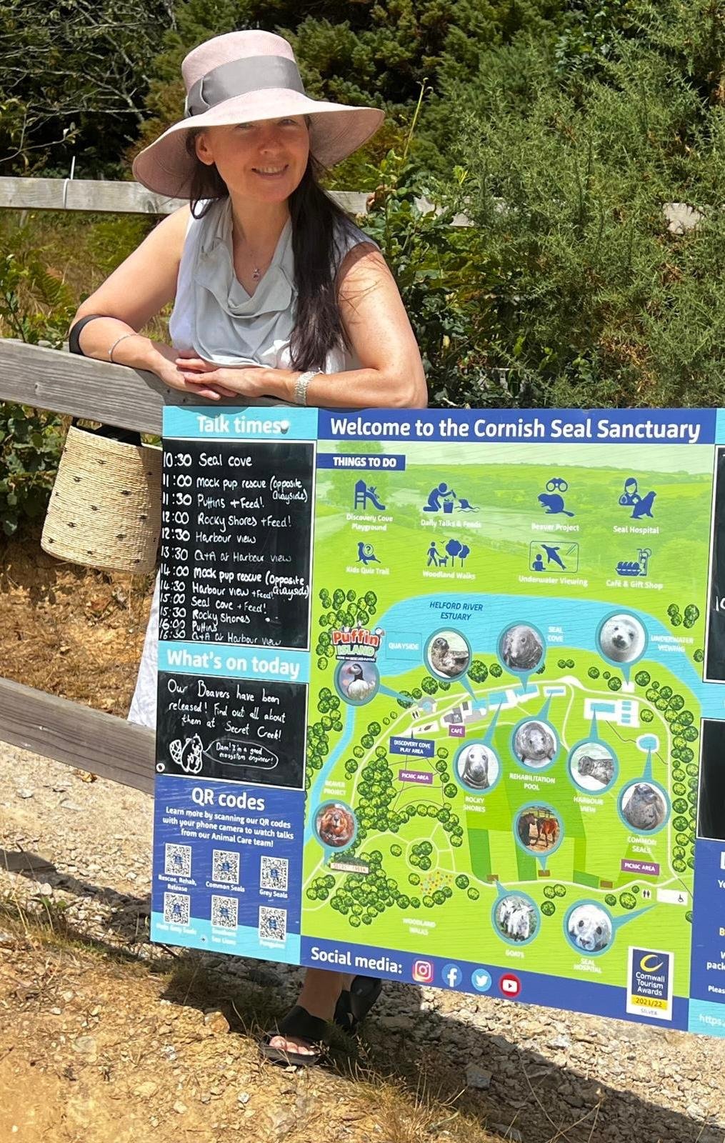 Woman smiling at Cornwall Seal Sanctuary, wearing a wide-brimmed hat and a sleeveless top, standing outdoors behind informational signs about the sanctuary's exhibits and activities.