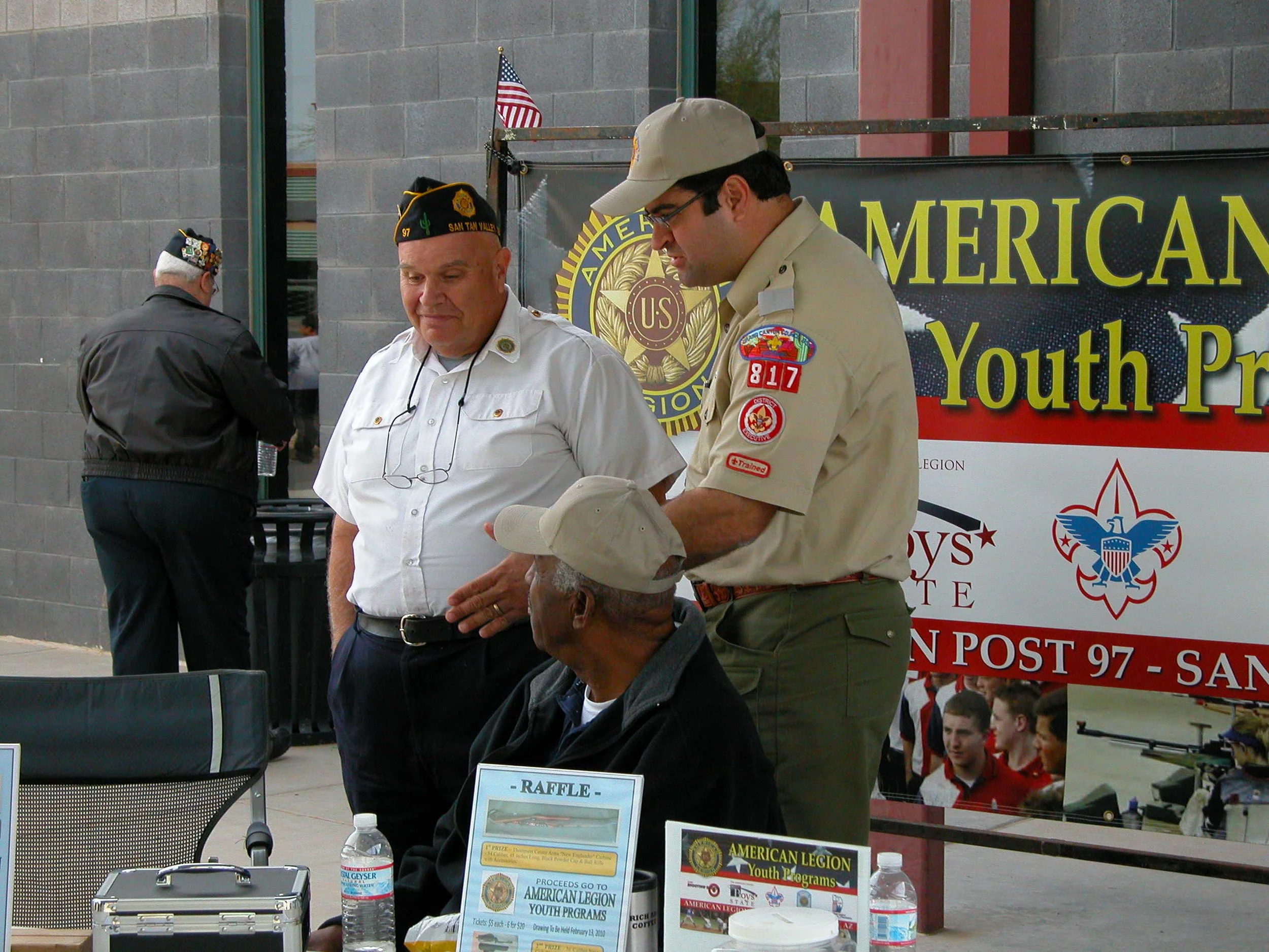 Three men in uniform at an outdoor event with a banner reading 'American Legion Youth Programs' and a display table with raffle info and bottles of water.