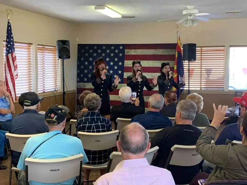 Three women in navy uniforms performing a song on stage with American and Arizona flags in the background.