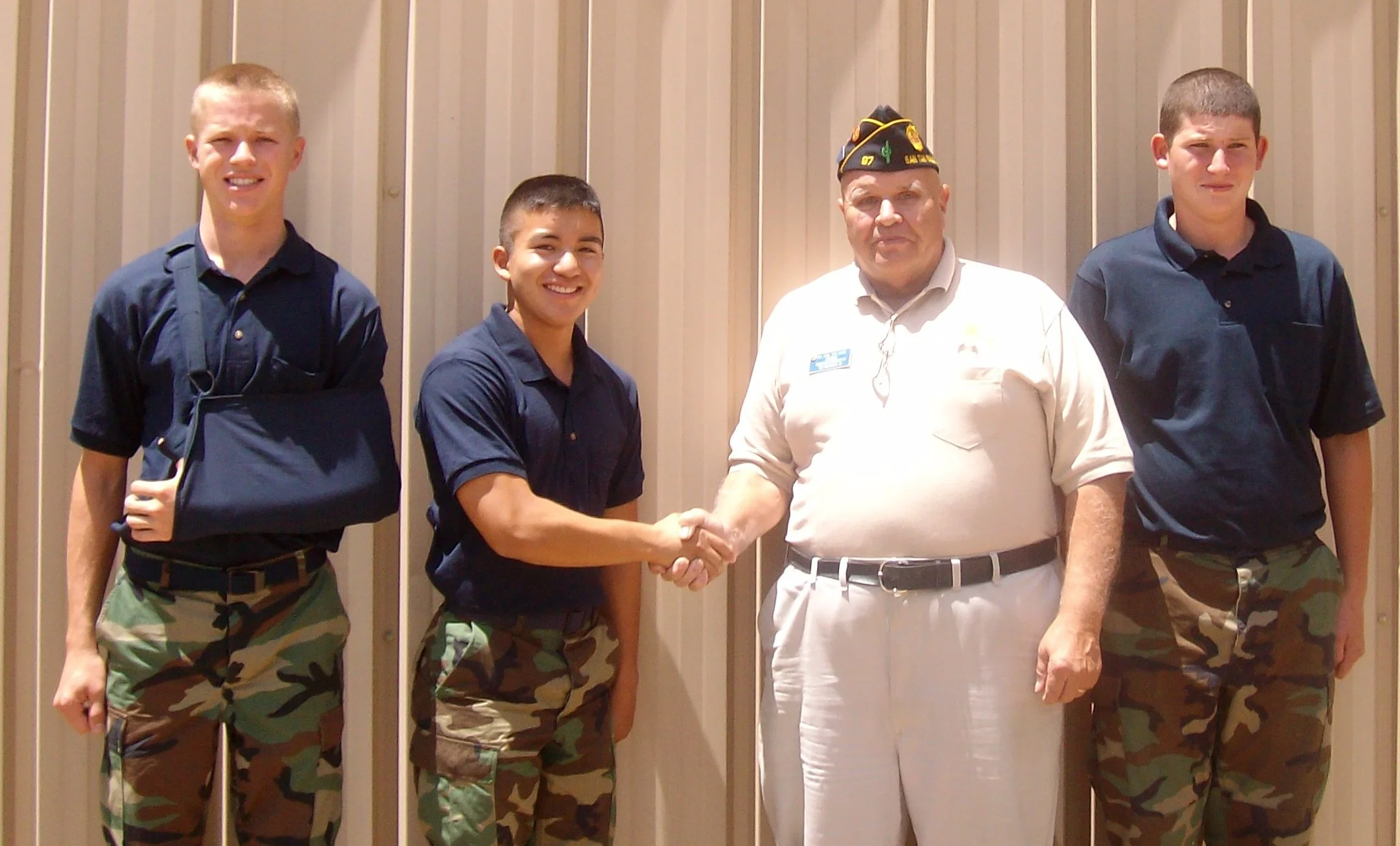 A young man in military camo pants and navy shirt shaking hands with an older man in a white uniform, with two other young men in military camo pants and navy shirts standing on either side. They are in front of a beige metal wall.