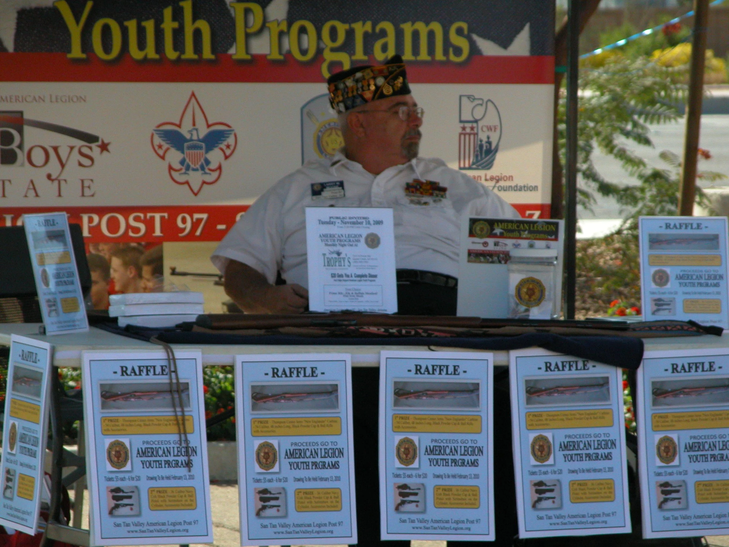 A man wearing military medals and a cap sits at a booth with flyers and signs for a raffle, promoting American Legion youth programs, with a large banner behind him reading 'Youth Programs' and multiple logos.
