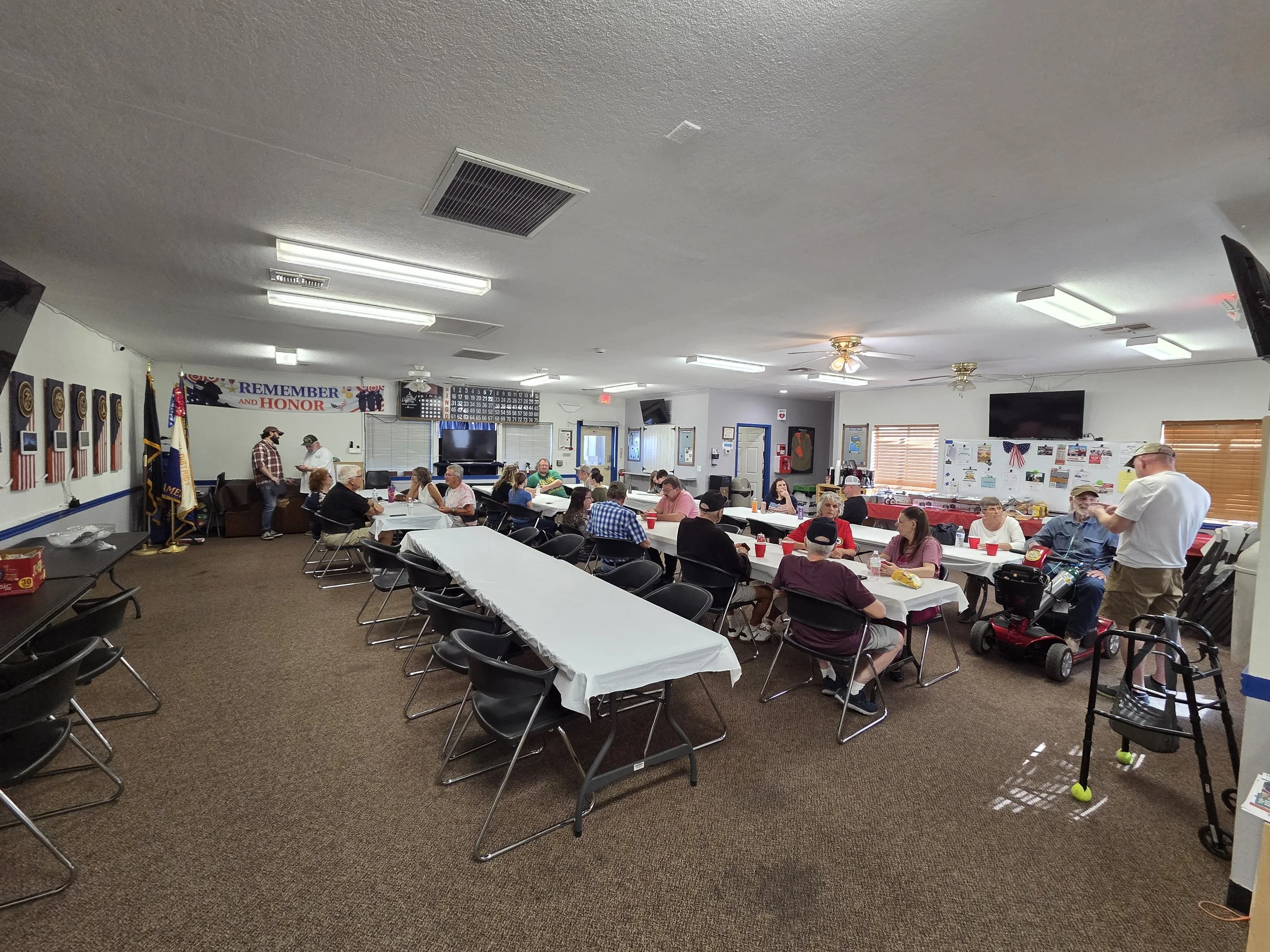 A community center or event hall filled with older adults sitting at tables covered with white tablecloths, engaged in conversations and activities. The room has a high ceiling with several fluorescent lights, ceiling fans, and patriotic decorations 