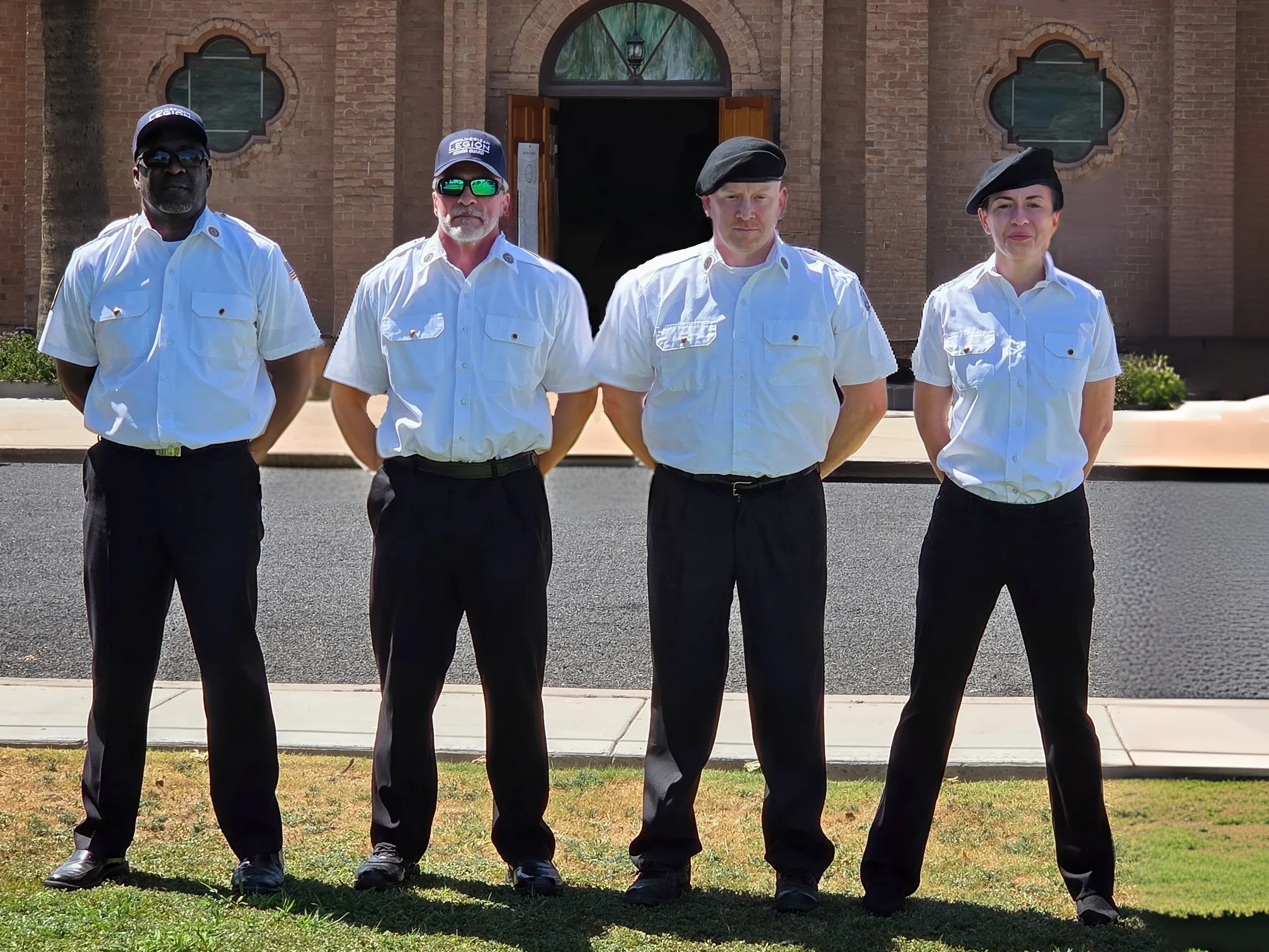 Four uniformed security personnel standing in front of a brick building with open wooden doors, wearing white shirts, dark pants, and dark berets or caps.