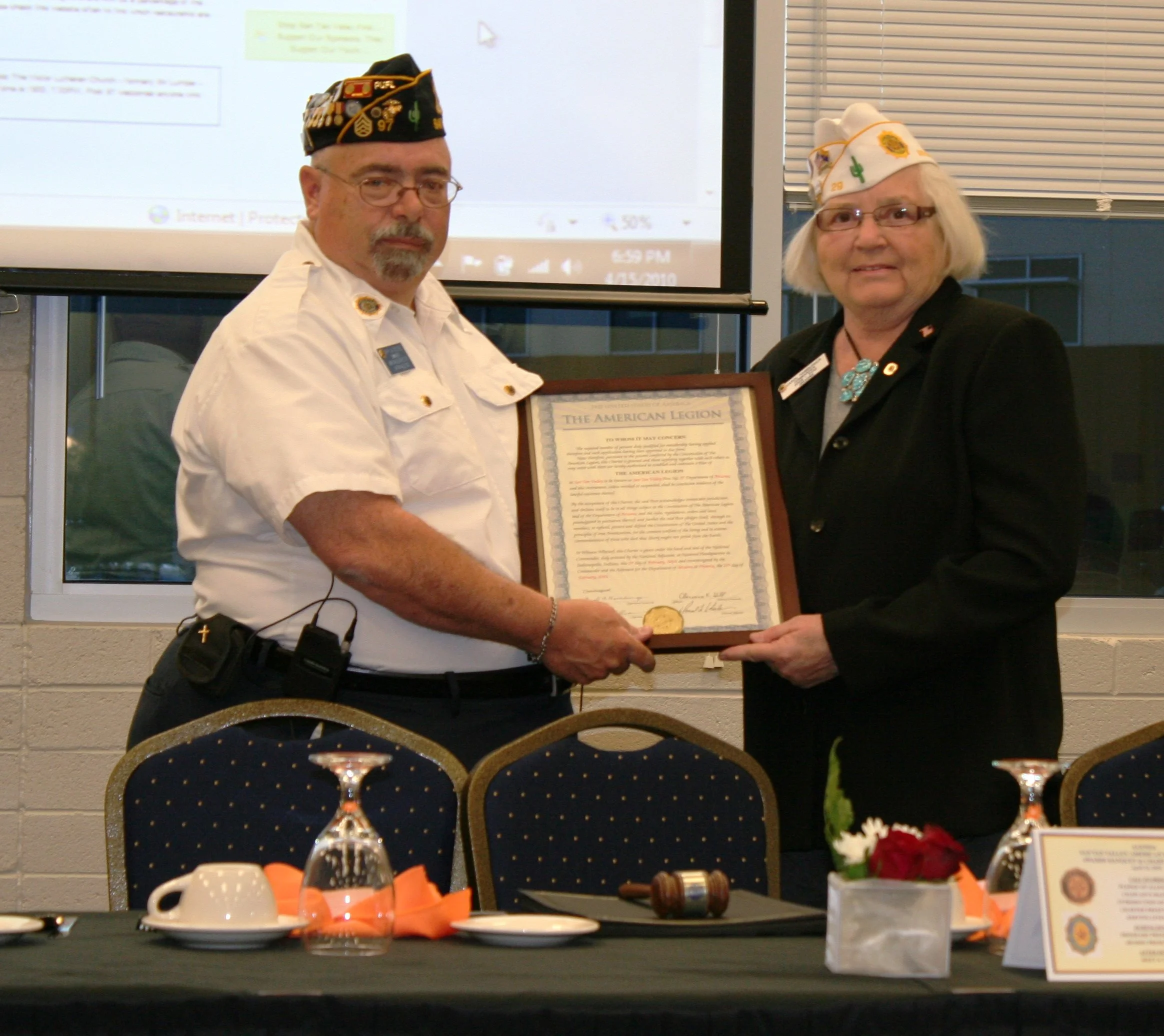 A man and a woman wearing veterans' organization hats, standing in front of a presentation screen, holding a framed certificate. The woman is smiling, and the man appears serious.