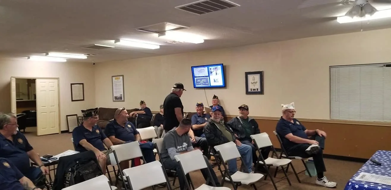 Group of veterans in a meeting room, sitting on white chairs, some wearing military service hats, with a man standing and talking.