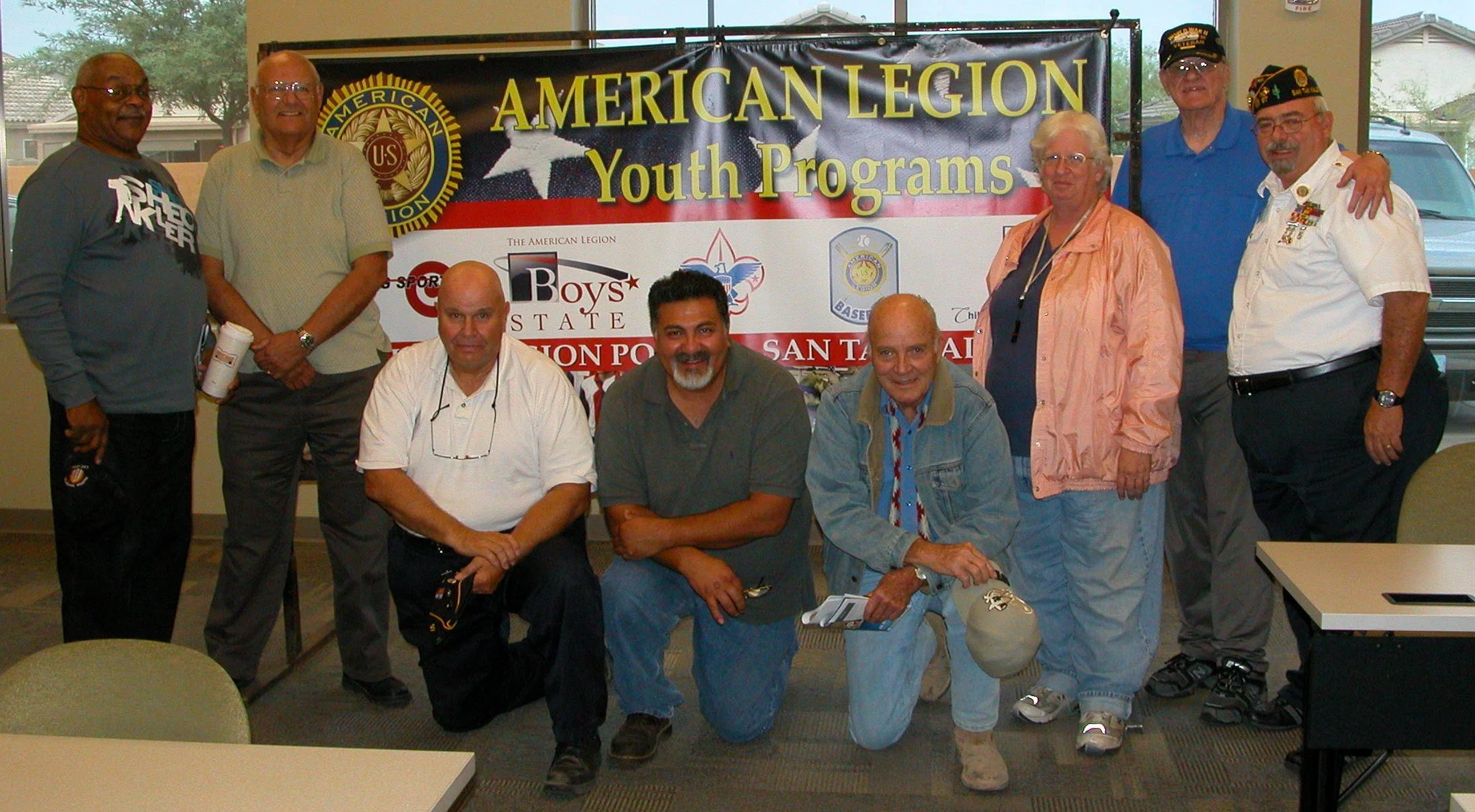 Group of diverse people posing in front of a banner that reads "American Legion Youth Programs" inside a room with windows showing trees and buildings outside.