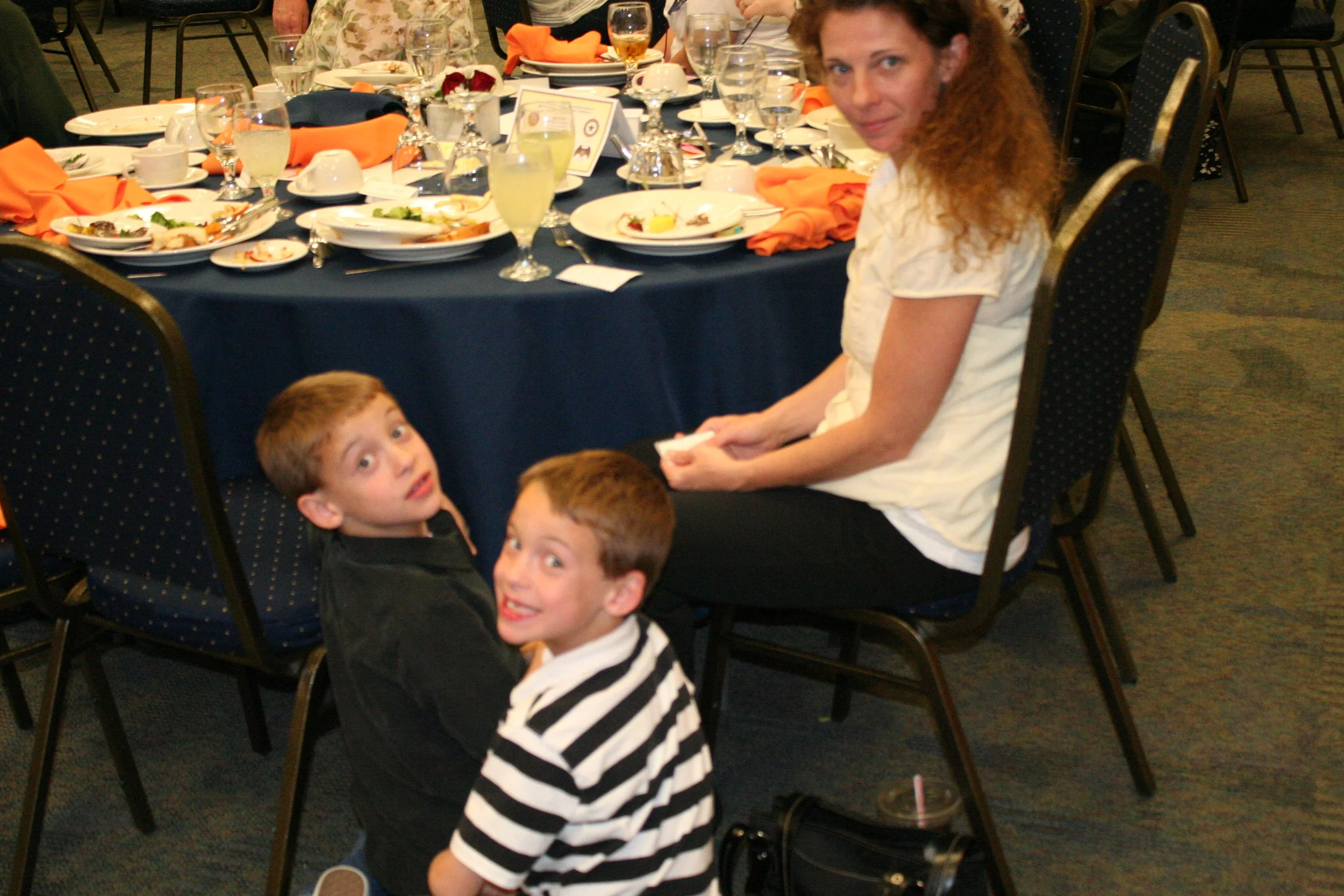 A woman with curly red hair and a white blouse sitting at a dinner table with four young boys. The table has dishes, glasses, and orange napkins, and is set for a meal at a formal event or gathering.