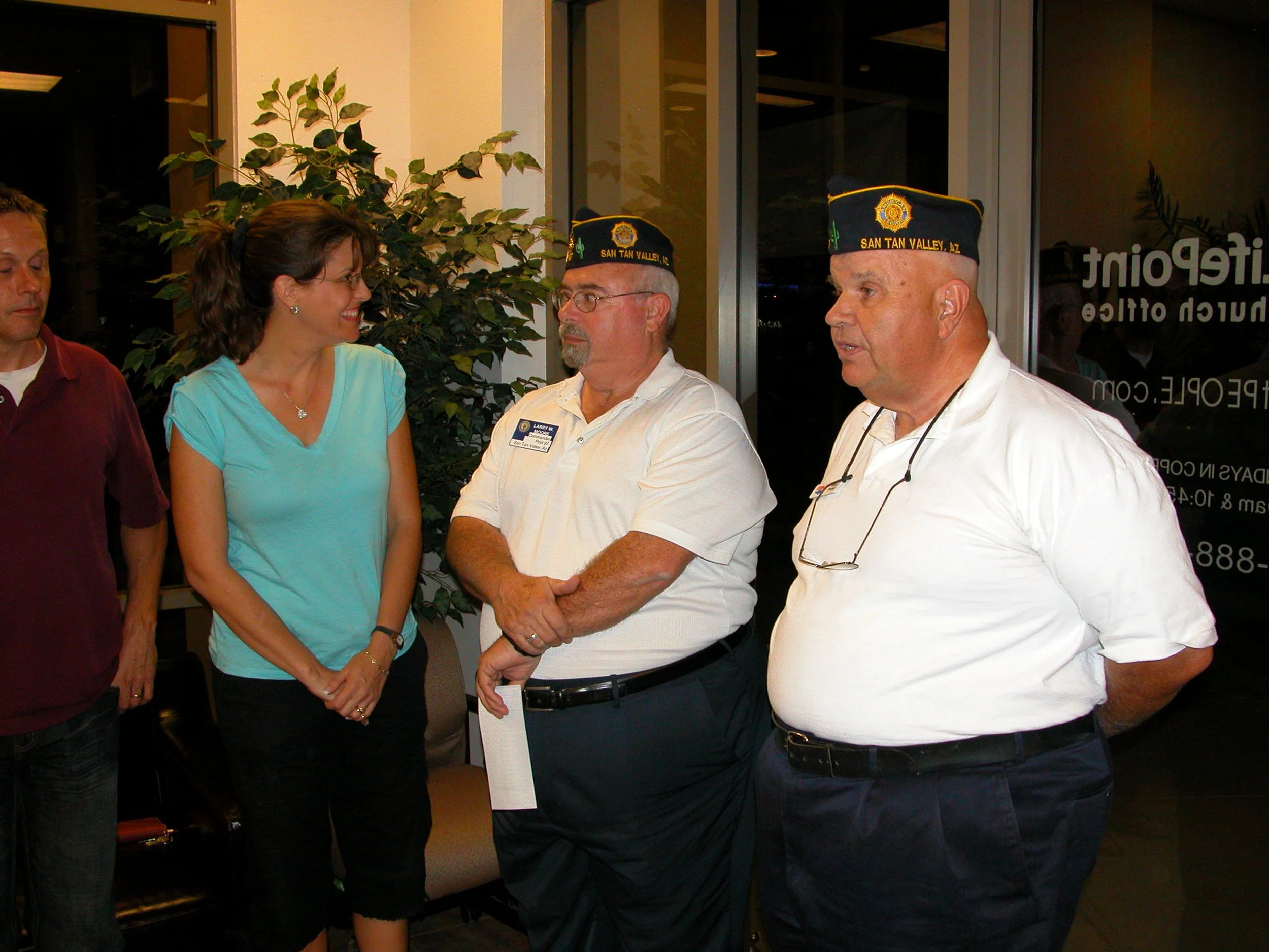 A group of five people standing inside a building, engaged in a conversation. Two men are wearing black caps with yellow trim and patches that read 'SAN TAN VALLEY, AZ'. The woman in a light blue shirt is smiling and talking to one of the men. The gr
