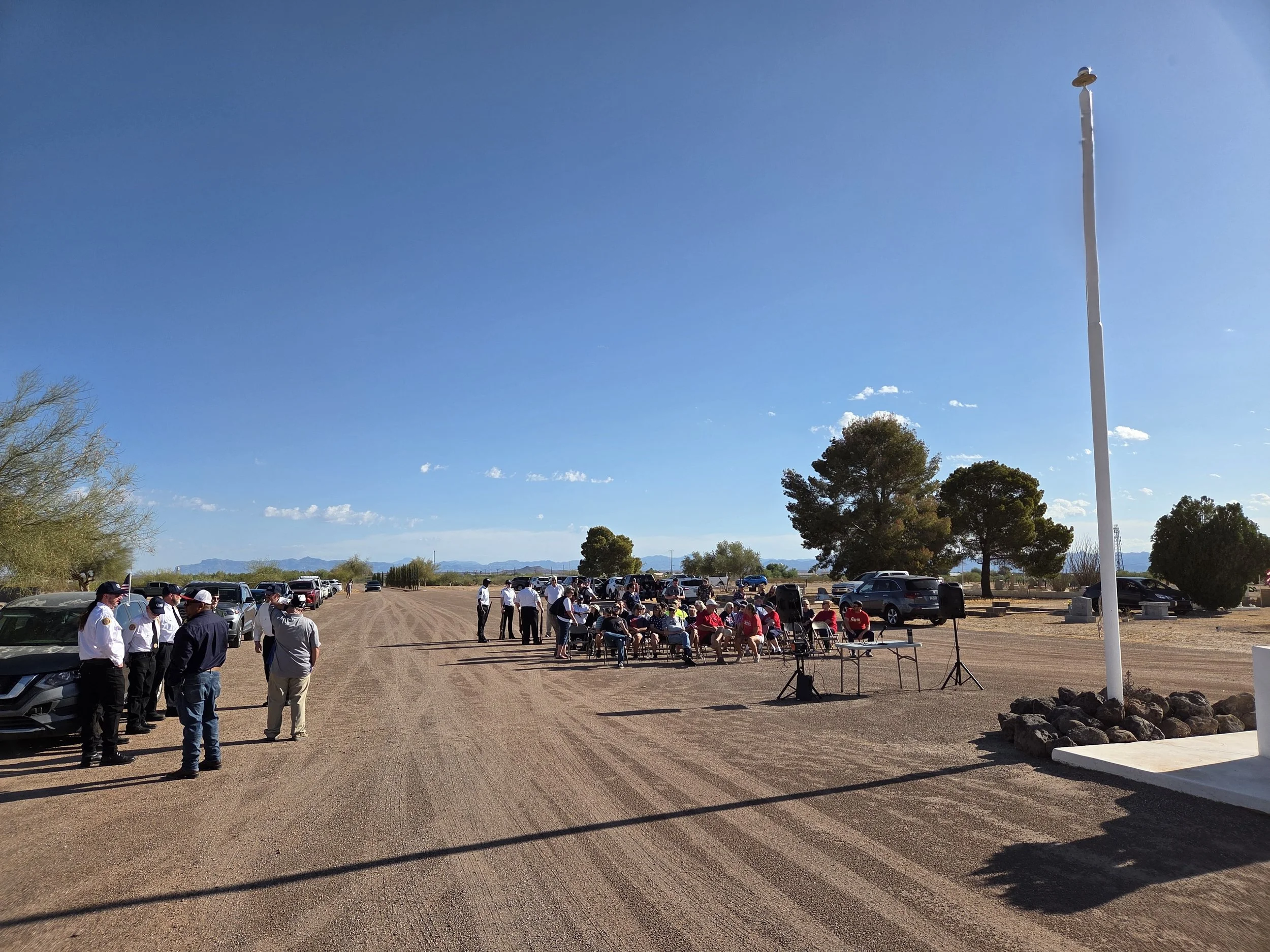 A group of people gathering outdoors on a sunny day with clear blue skies, some seated and some standing, near a gravel parking lot with parked cars, trees, and mountains in the background.