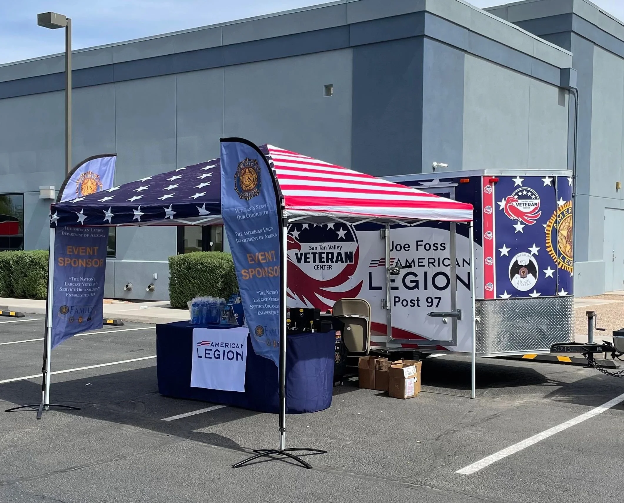 A parking lot setup with a tent and a trailer for a veteran event. The tent has patriotic flags and banners, including one that says 'Event Sponsor' and features the American Legion emblem. The trailer displays a logo for the San Tan Valley American 