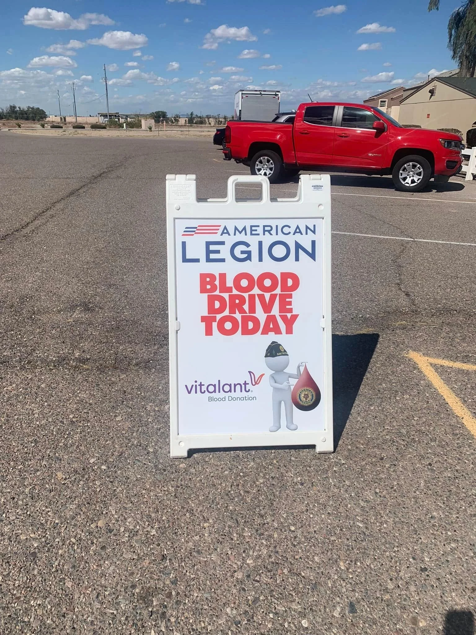 A sign in a parking lot for a blood donation event organized by American Legion, with the words "Blood Drive Today" and the logo of Vitalant. In the background, a red truck is parked with a small building and blue sky with clouds above.