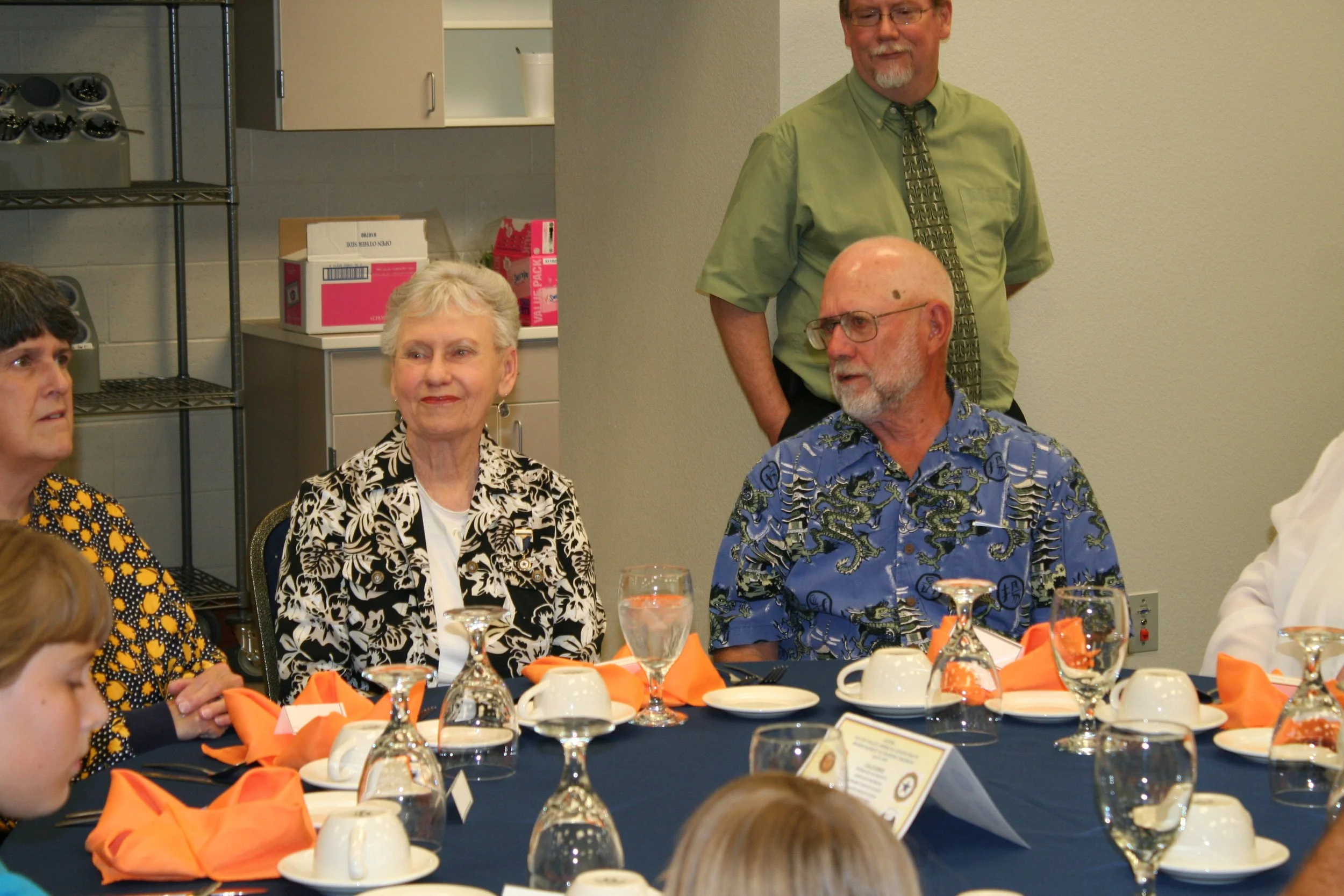 People sitting around a dining table at a formal event, with place settings including glasses, plates, and orange napkins, in a room with a kitchen in the background.