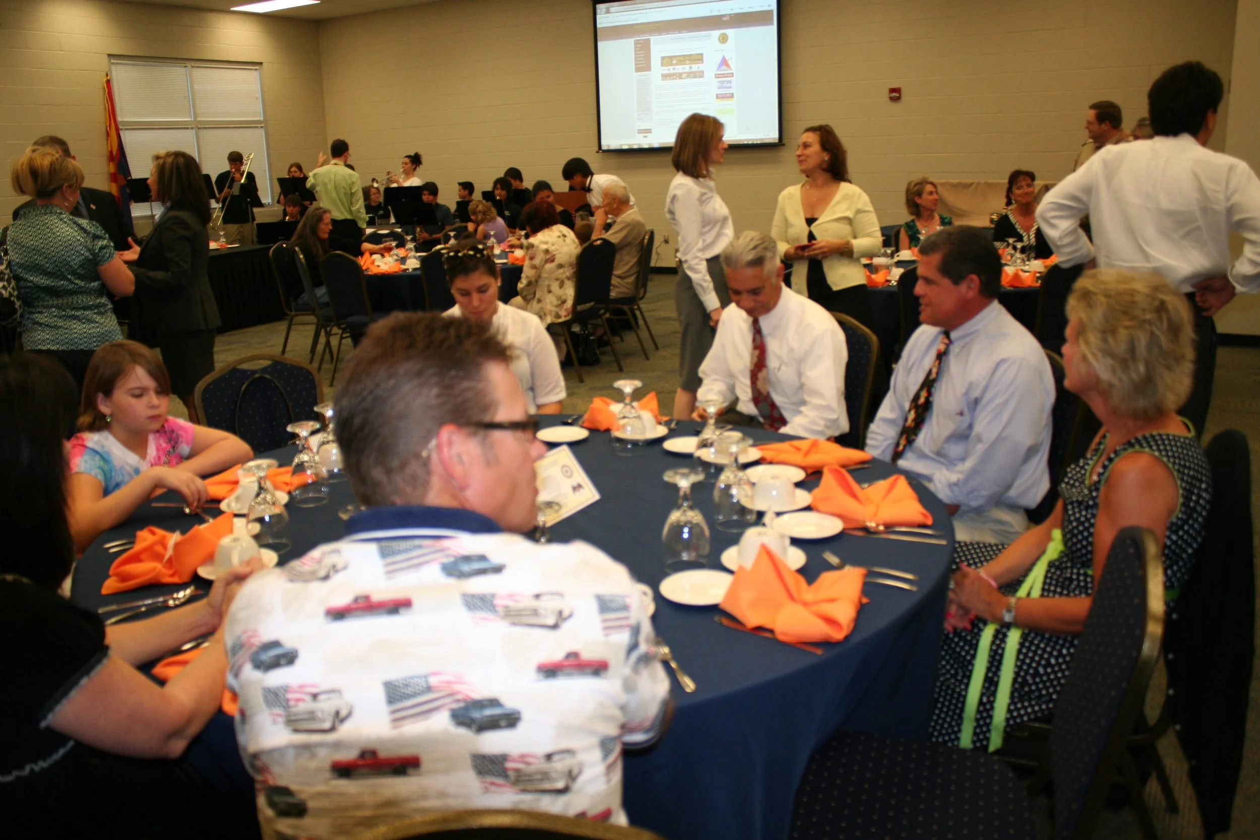 Guests seated around a banquet table with orange napkins at a formal event, with some people standing and talking, a band playing in the background, and a large screen displaying a presentation.