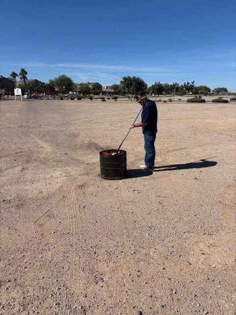 A man in a hat and sunglasses tending to a fire in a metal drum in an empty sandy lot under a clear blue sky.