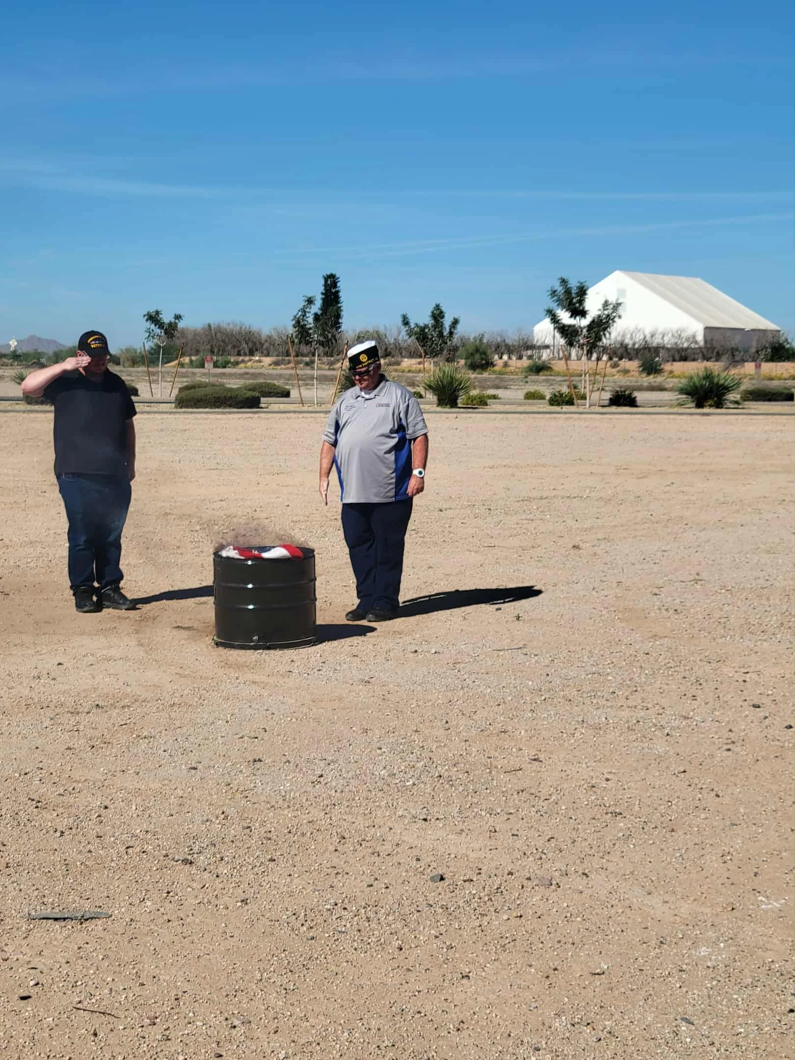 Two men in military uniform hats standing outdoors near a black barrel with a red and white object on top, in a desert-like area with sparse trees and bushes, and a large white tent in the background under a blue sky.