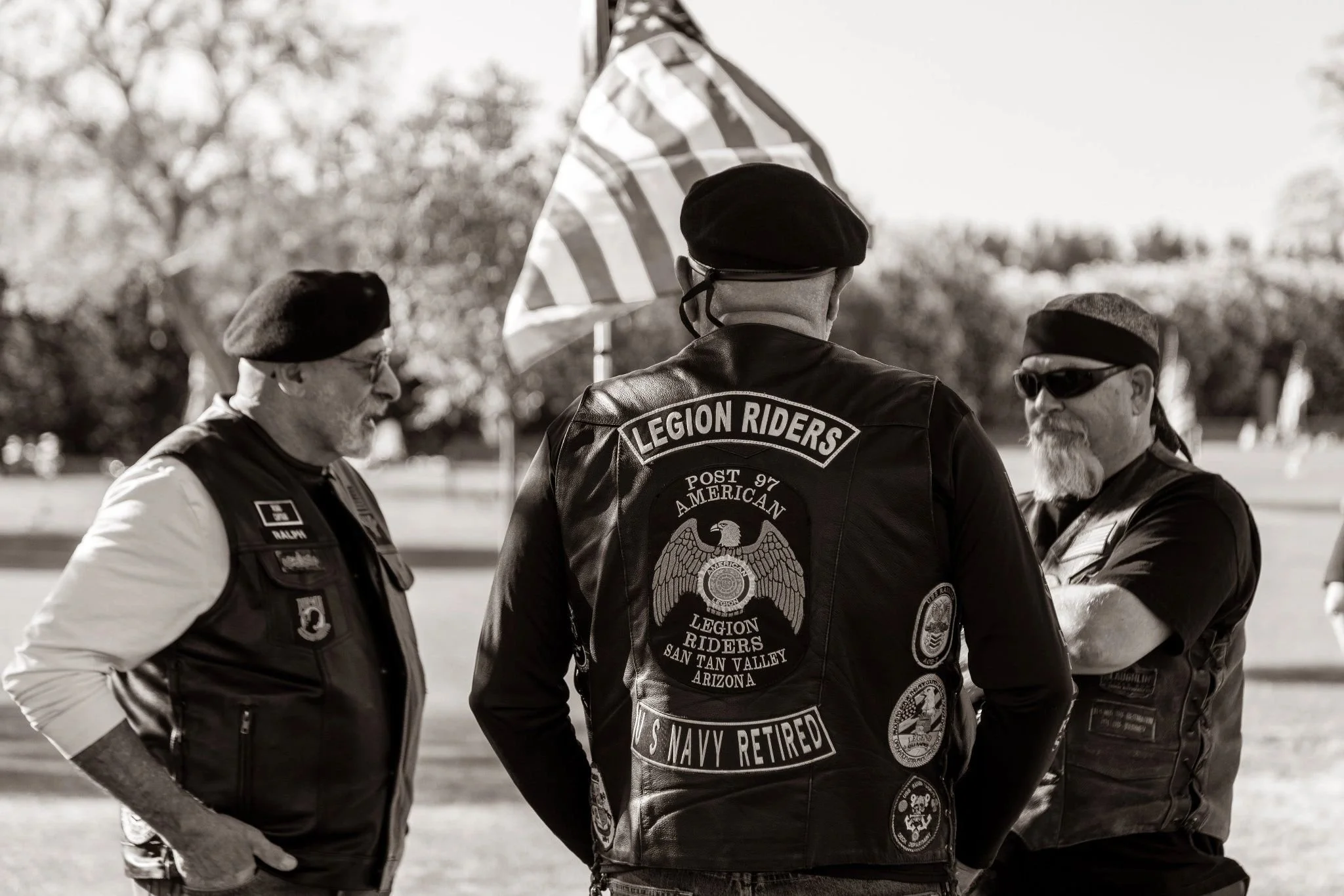 Group of four men wearing black leather jackets with patches, gathered outdoors, one wearing a beret, two with sunglasses, one with a flat cap, discussing with American flags in the background.