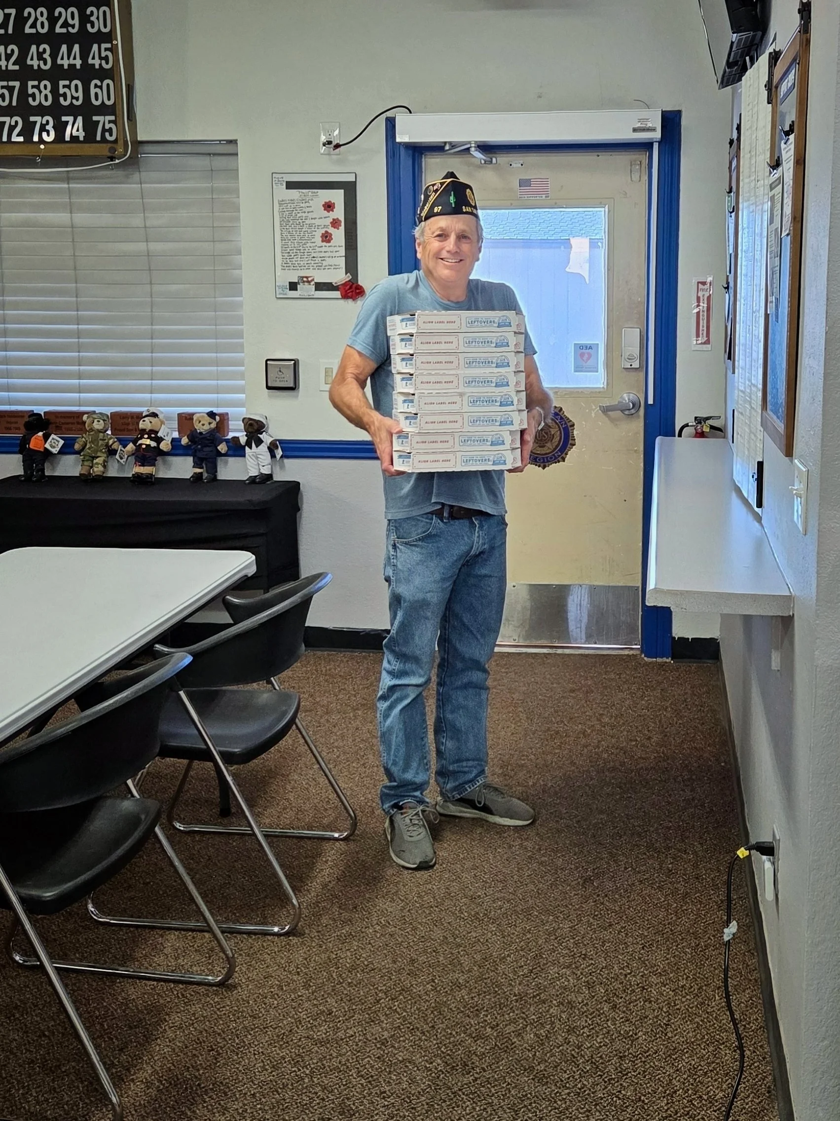 Older man wearing a black veteran's cap and a blue T-shirt, smiling while holding six pizza boxes stacked in a vending style at a community center or veteran hall with couches, teddy bear dolls, and informational posters on the wall.