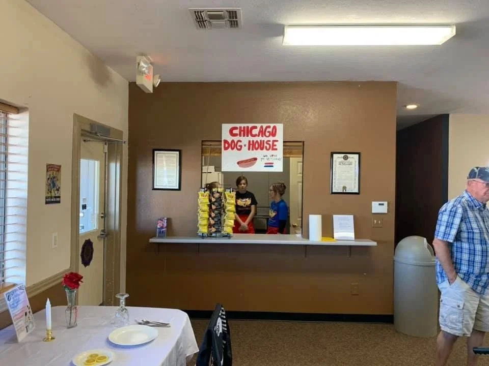Interior of a restaurant with a counter for ordering food. There are two women working behind the counter and a man standing to the right. A sign reads 'Chicago Dog House'. On the table in the foreground, there are plates, utensils, and a small flowe
