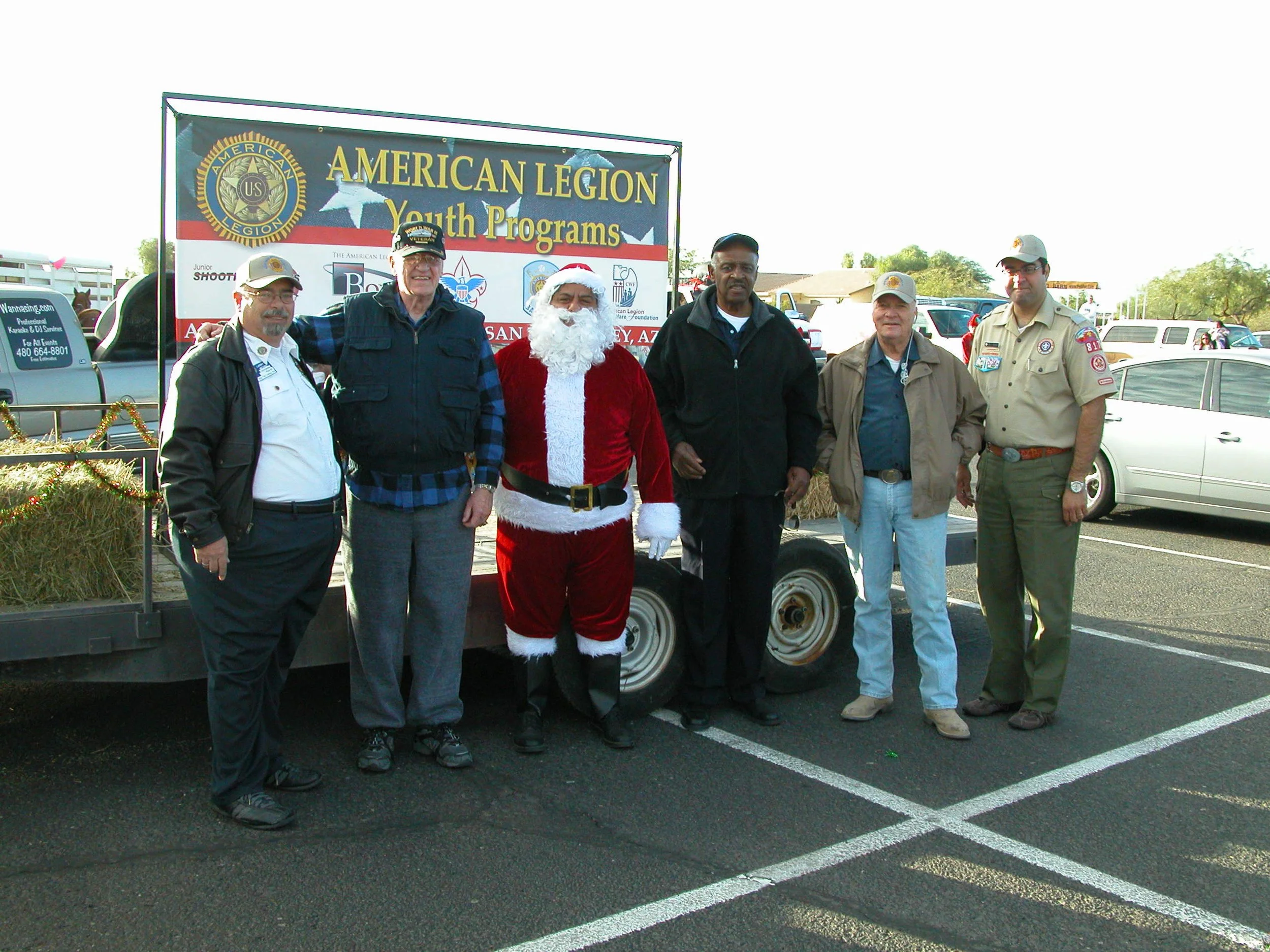 Group of six men standing together outdoors in front of a large sign for American Legion Youth Programs. One man is dressed as Santa Claus. The setting appears to be a parking lot with cars and a building in the background.