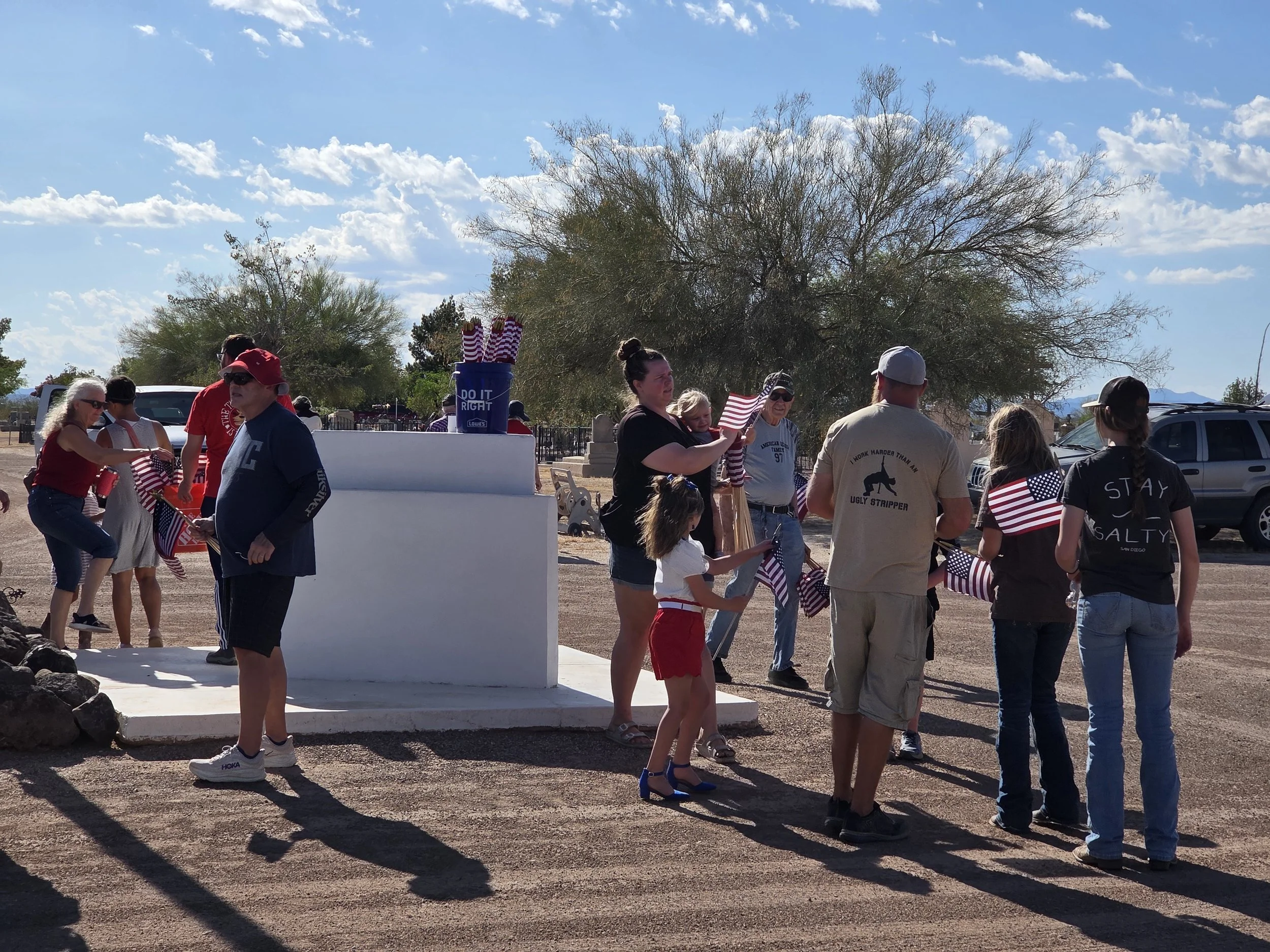 A group of people gathered outdoors under a sunny sky with scattered clouds. Some are holding American flags and appear to be participating in a patriotic event. The background includes trees, parked cars, and a distant mountain range.