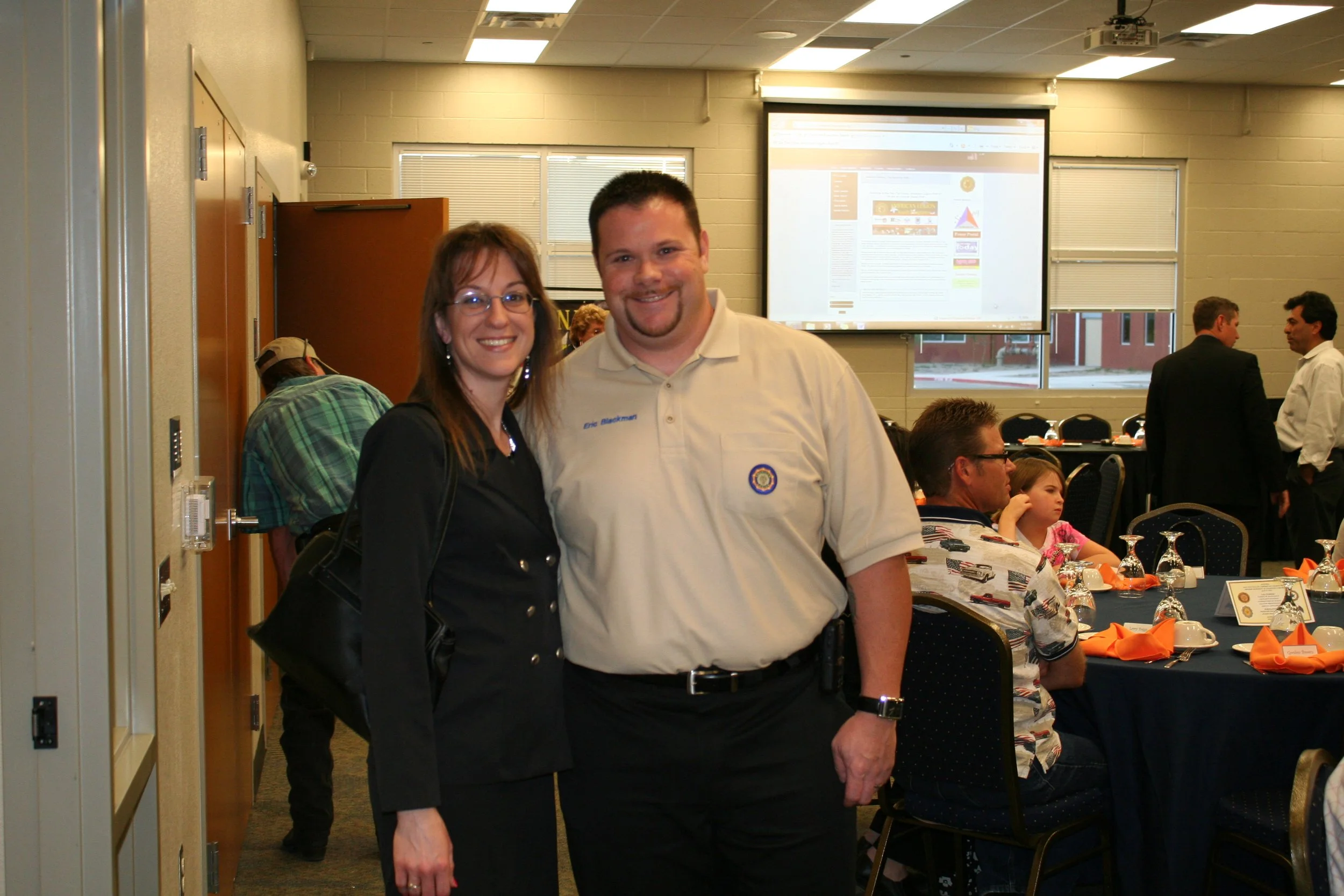 Two people smiling at a gathering or conference, with tables decorated with orange napkins and glassware, and a large screen displaying a website or presentation behind them.