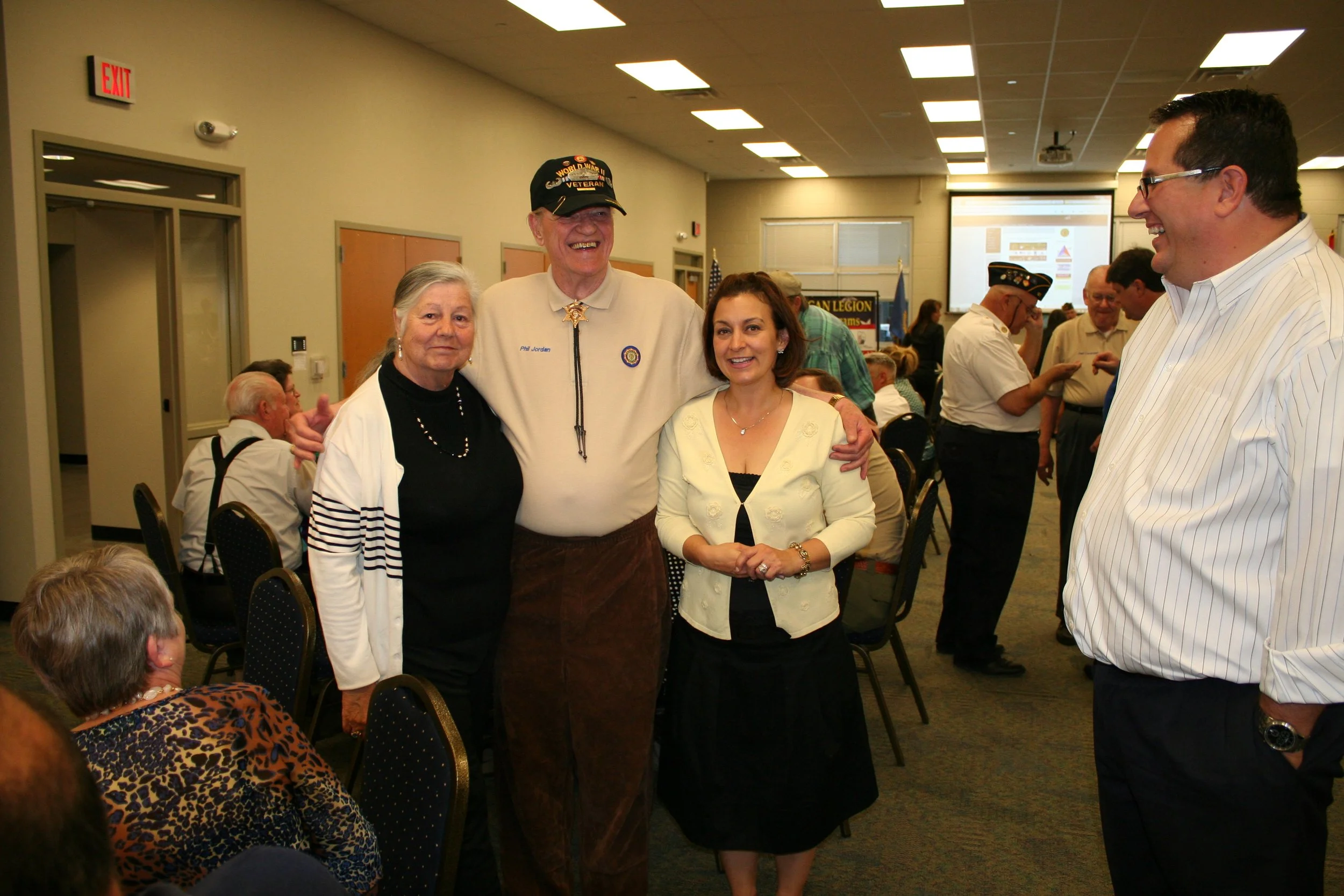 Group photo of four people smiling at an indoor event, with a large screen in the background and other attendees mingling, some wearing hats.