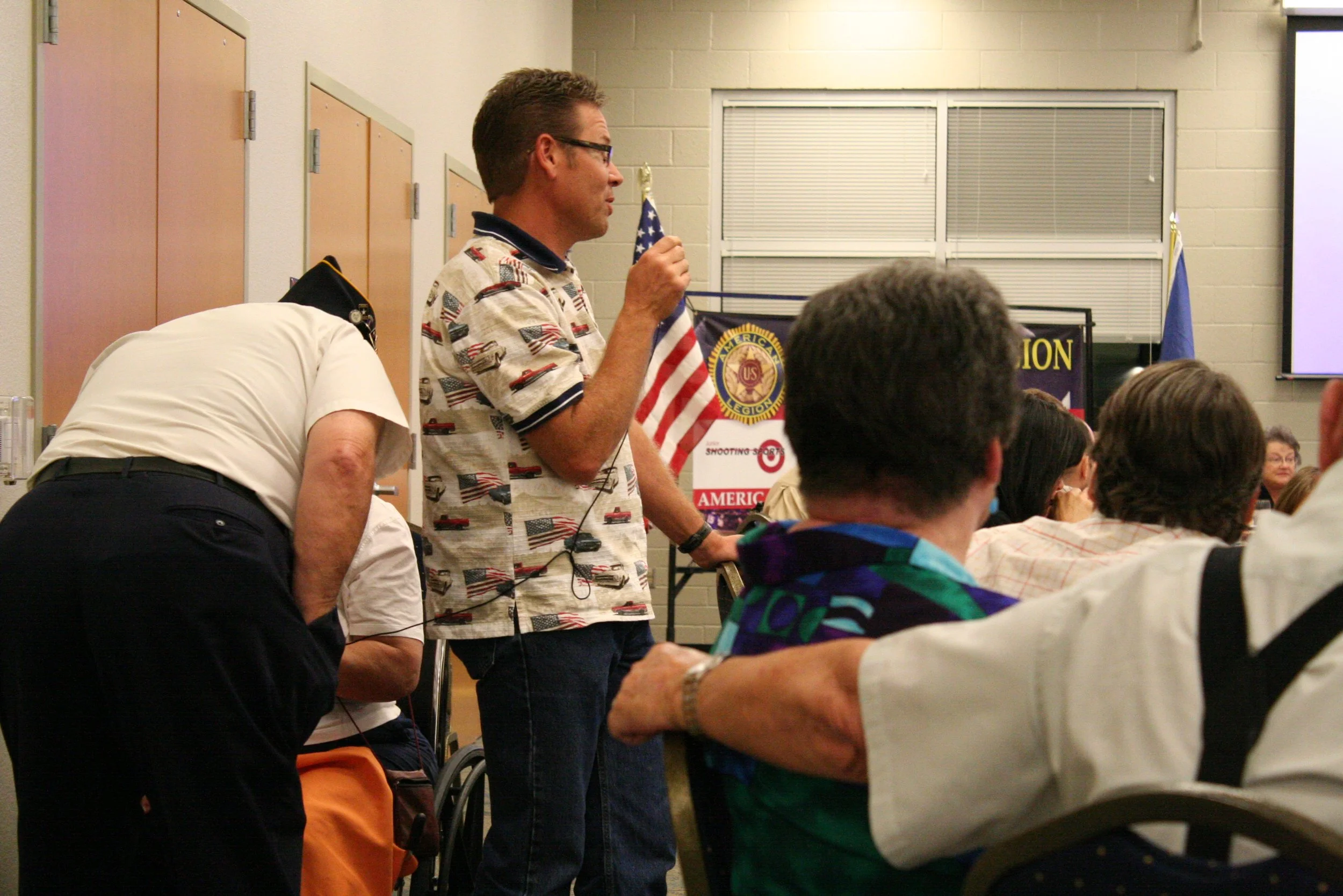 A man wearing glasses and a shirt with American flags is holding an American flag in a classroom or conference room, speaking to an audience.