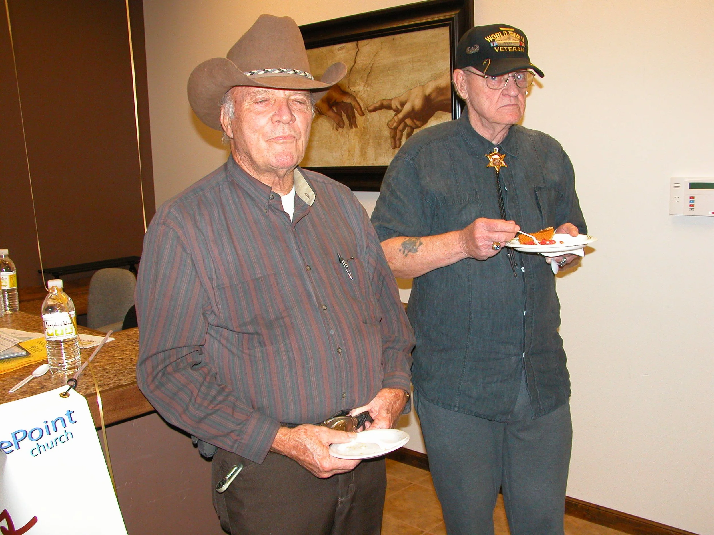 Two elderly men standing indoors, one wearing a cowboy hat and the other a black cap with military insignia, both holding small plates. The man in the cowboy hat has his eyes closed and is smiling slightly. Behind them, a framed artwork depicting han