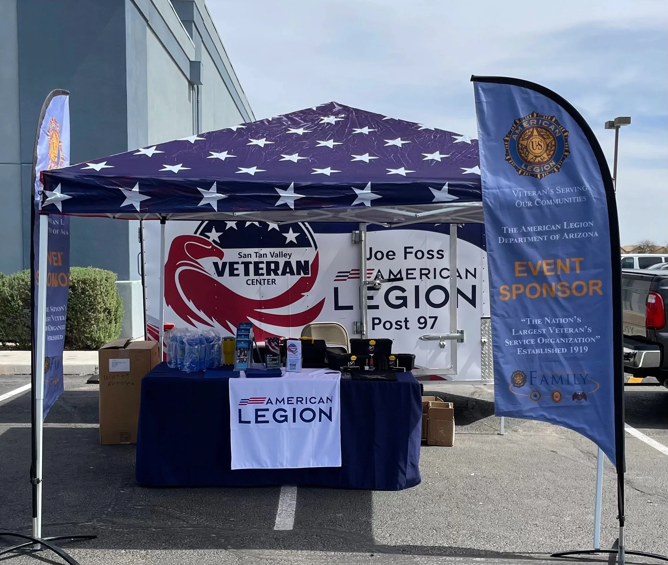 A tent with a blue star-spangled American flag pattern canopy set up at an outdoor event, displaying banners and signs for the American Legion, with additional flags and promotional materials underneath.